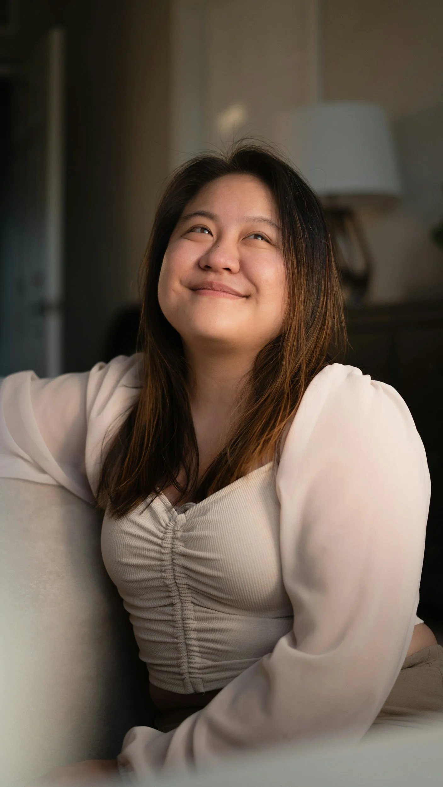 A woman with long dark hair and a light-colored blouse smiling and looking upward in a cozy indoor setting.