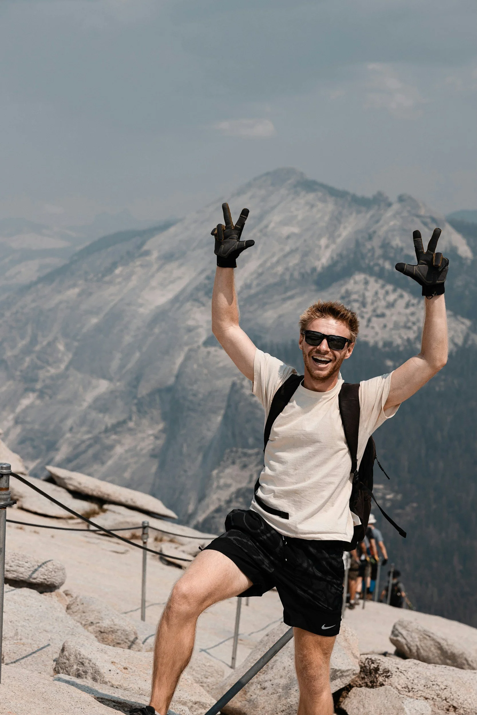 A smiling man wearing sunglasses and a white t-shirt making peace signs with both hands. He is standing outdoors in front of mountains with cloudy skies. He has a black backpack and black shorts, with one leg bent and the other straight. There are some rocks and a railing in the foreground.