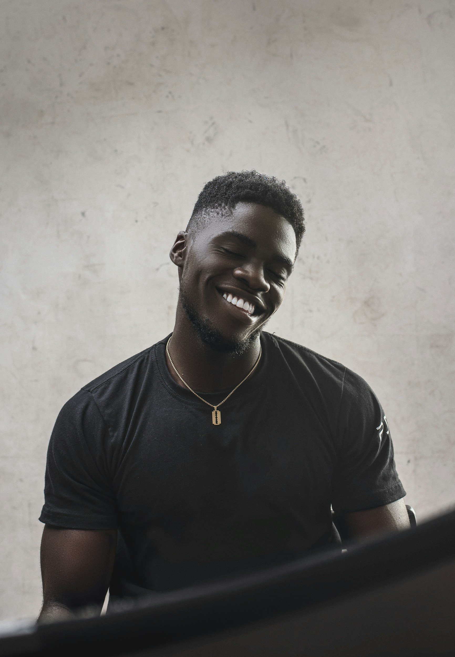 Young man with dark skin, short curly hair, smiling with eyes closed, wearing a black t-shirt, a gold necklace, and sitting in front of a plain light-colored wall.