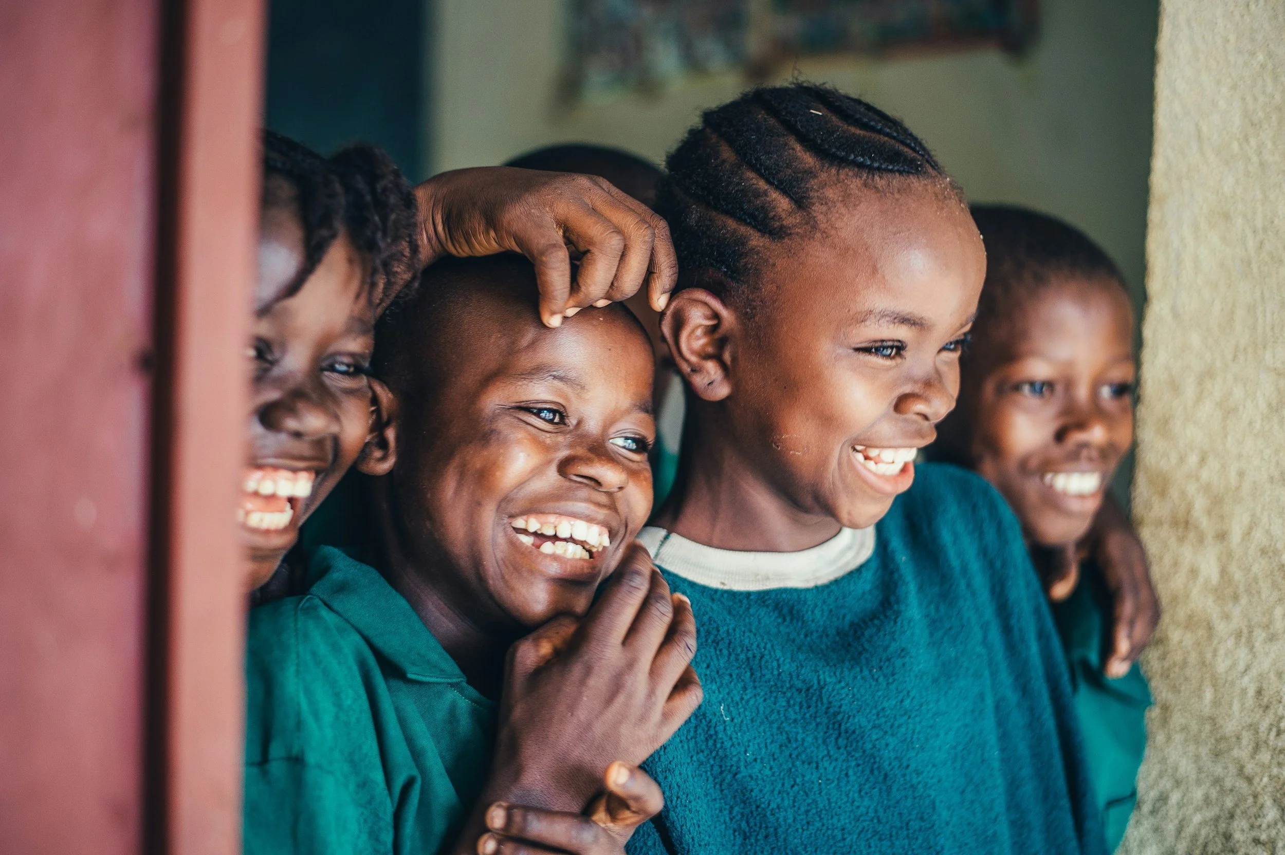 Children with smiles and laughter, looking out from behind a wall, possibly enjoying a moment of play or discovery.