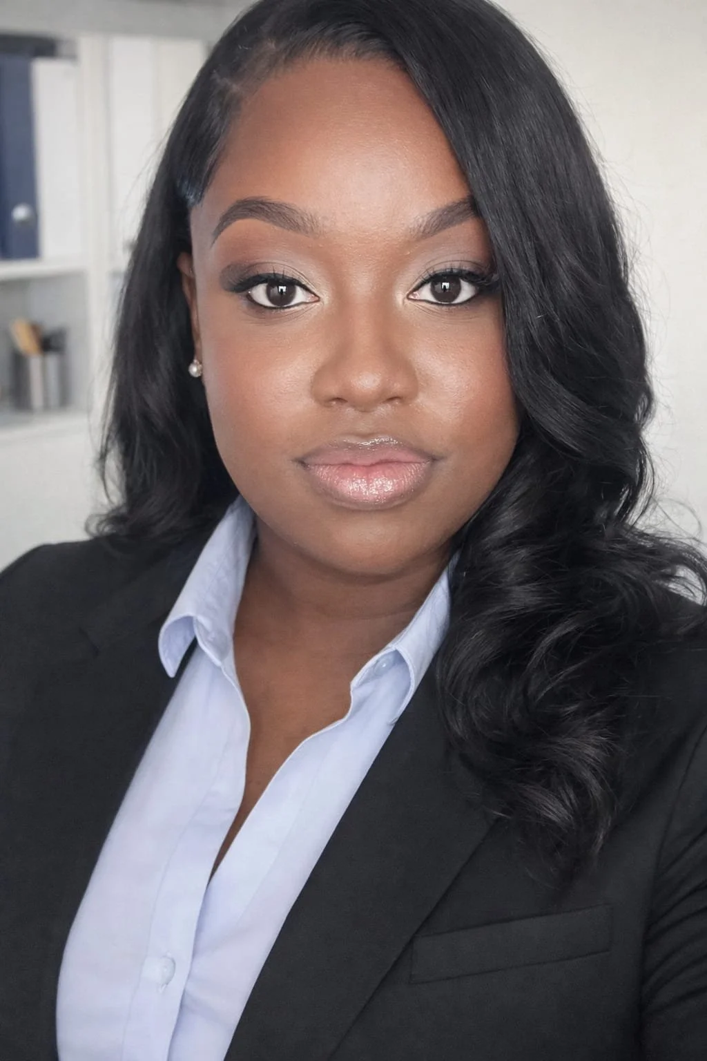A professional woman with dark hair and makeup, wearing a black blazer and a white collared shirt, in an office setting.