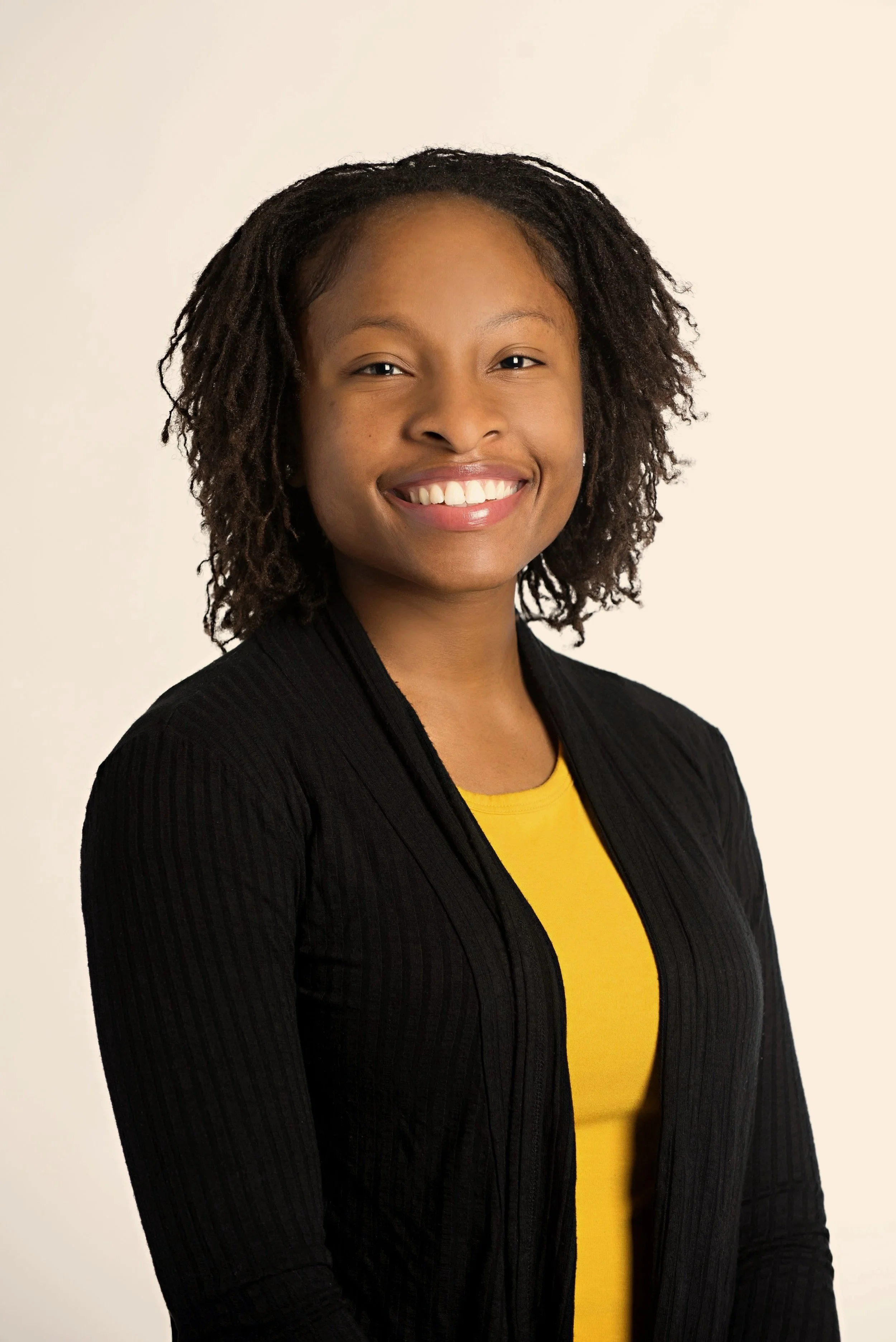 A smiling woman with curly hair wearing a black jacket over a yellow top against a plain background.