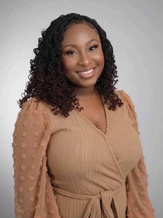 A woman with curly black hair smiling at the camera, wearing a beige top with textured sleeves tied at the waist.