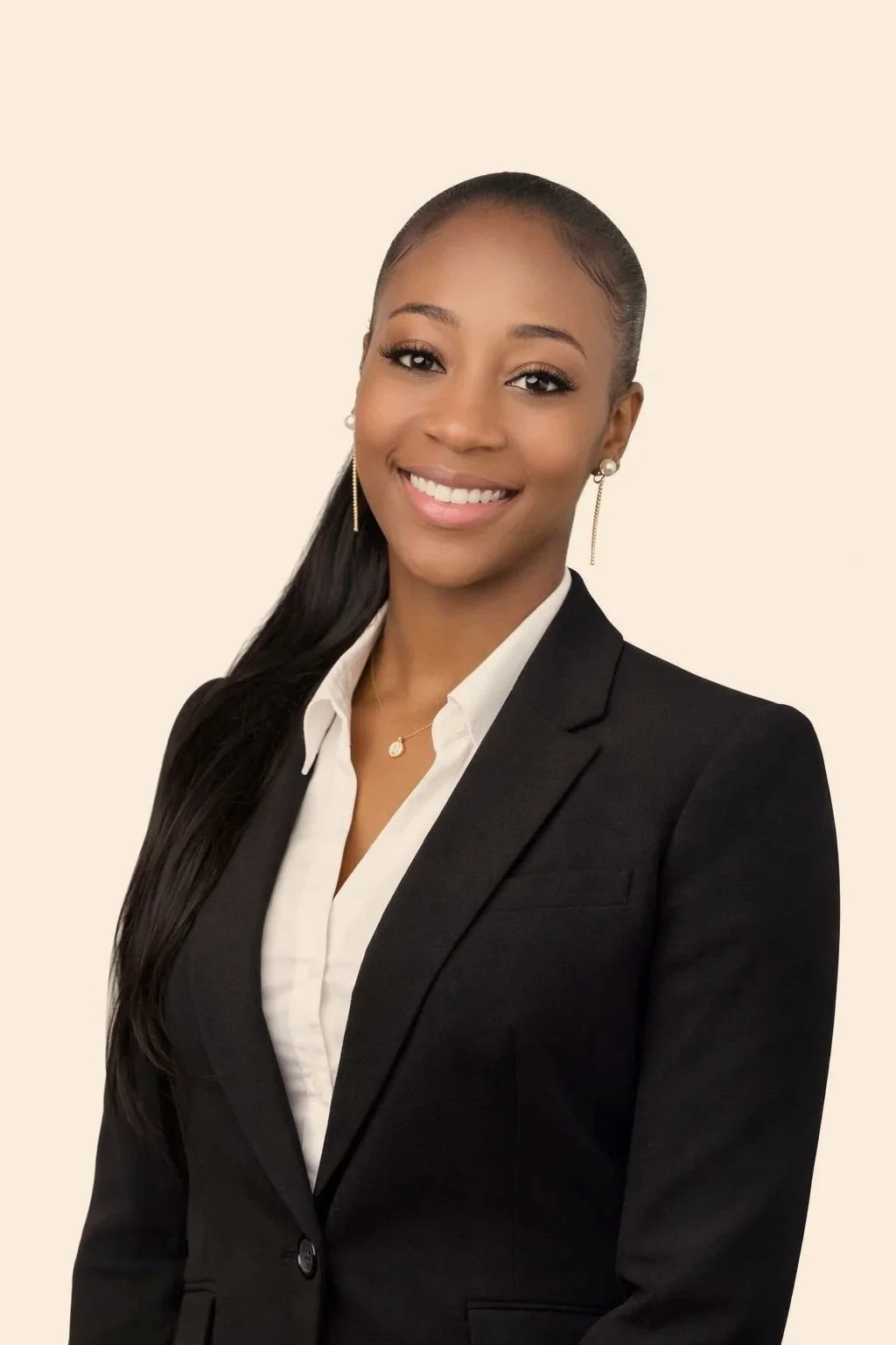 Portrait of a young woman in a black blazer and white shirt, smiling, with long dark hair, earrings, jewelry, and a plain light background.