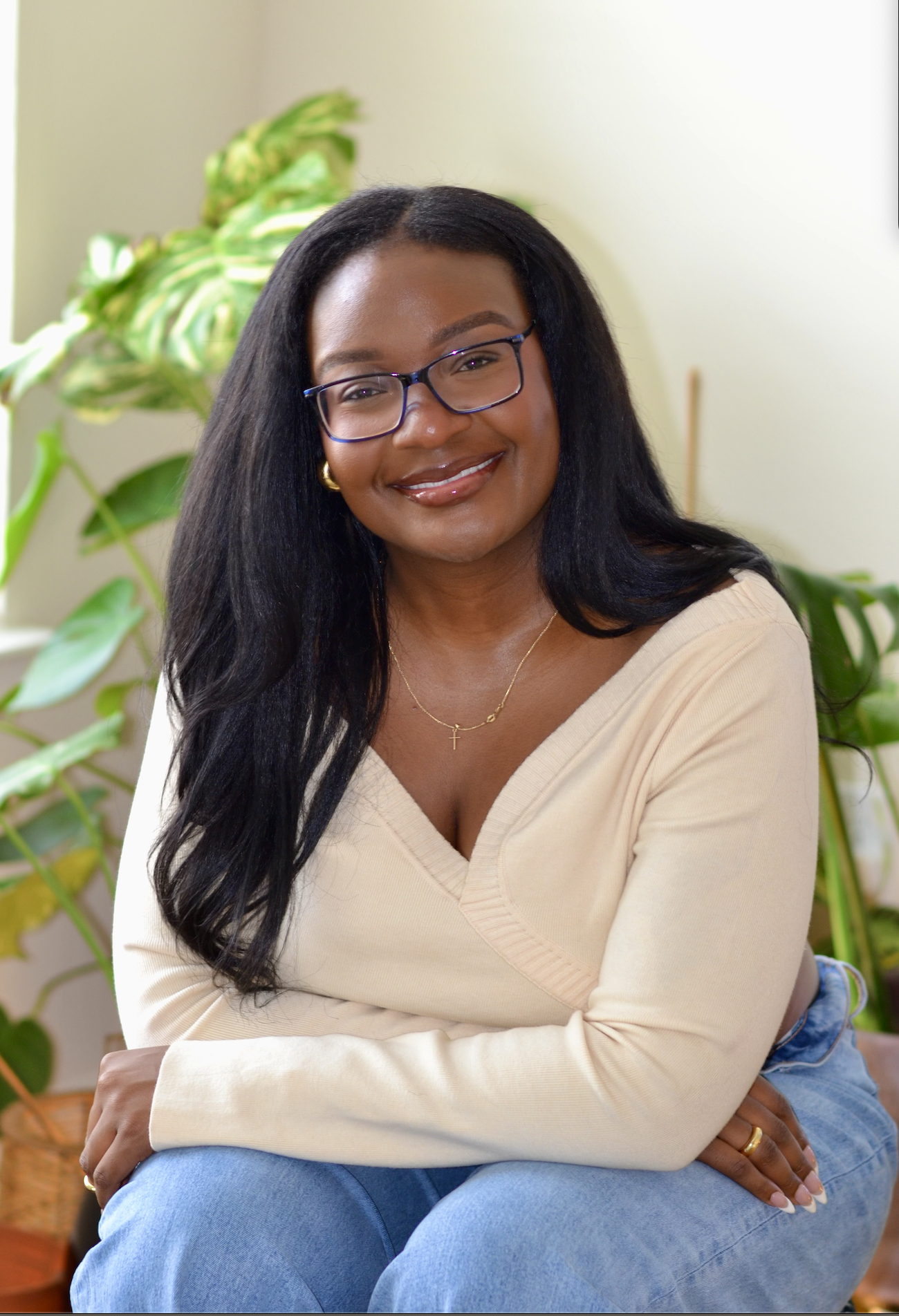 Smiling woman with black hair, glasses, and jewelry, sitting in a room with green plants.