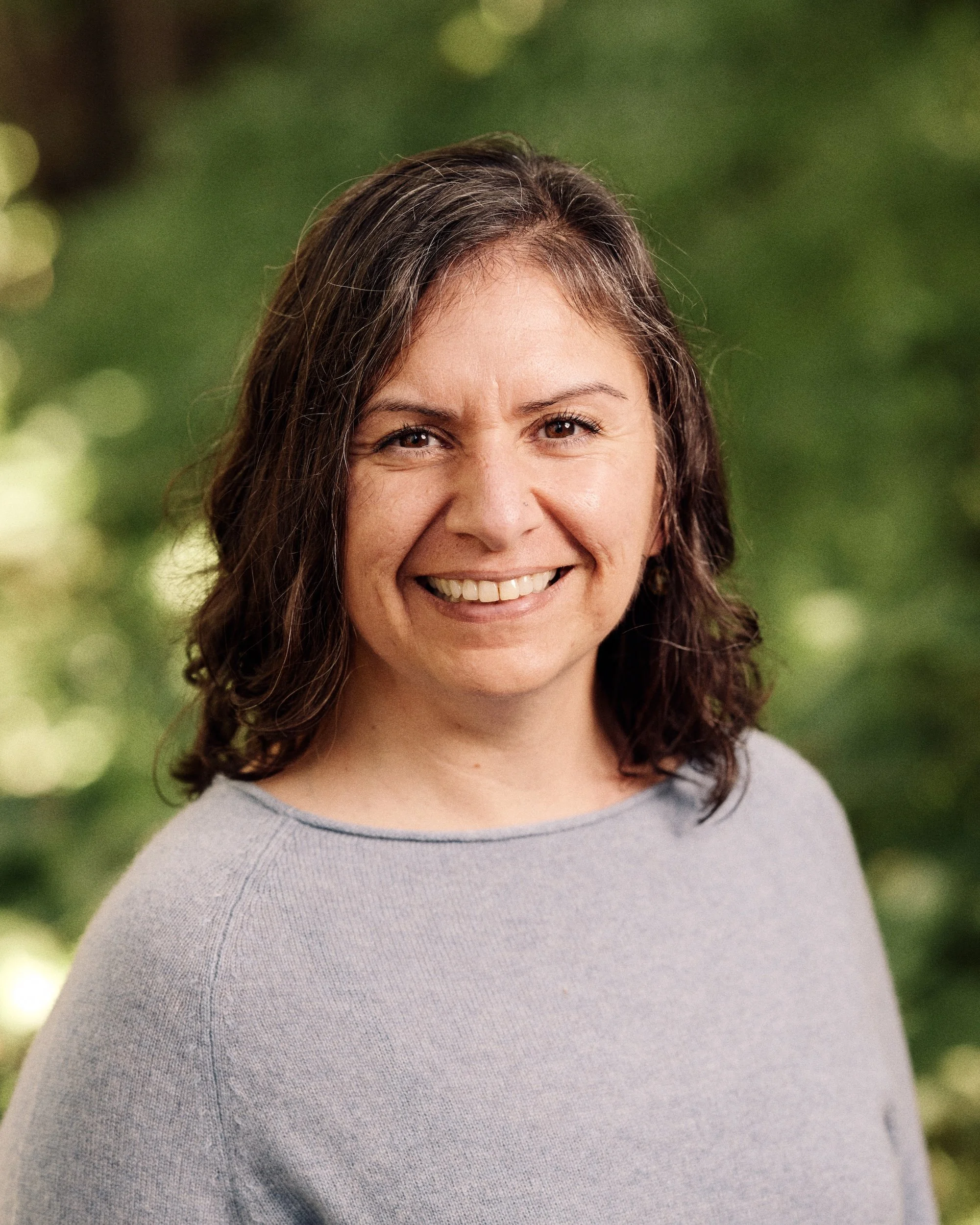 Close-up of a woman smiling, wearing a light gray top, with green blurred background.