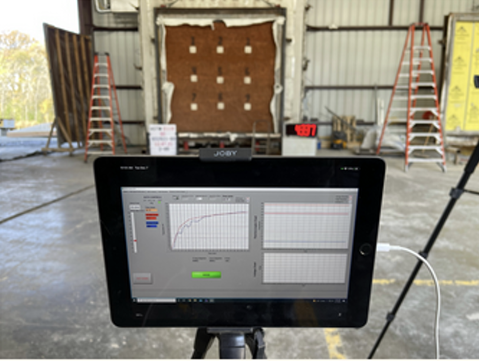 A construction or testing site inside a building with two orange ladders, a wall with foam insulation, and a digital display showing data. An electronic device on a tripod captures the scene.