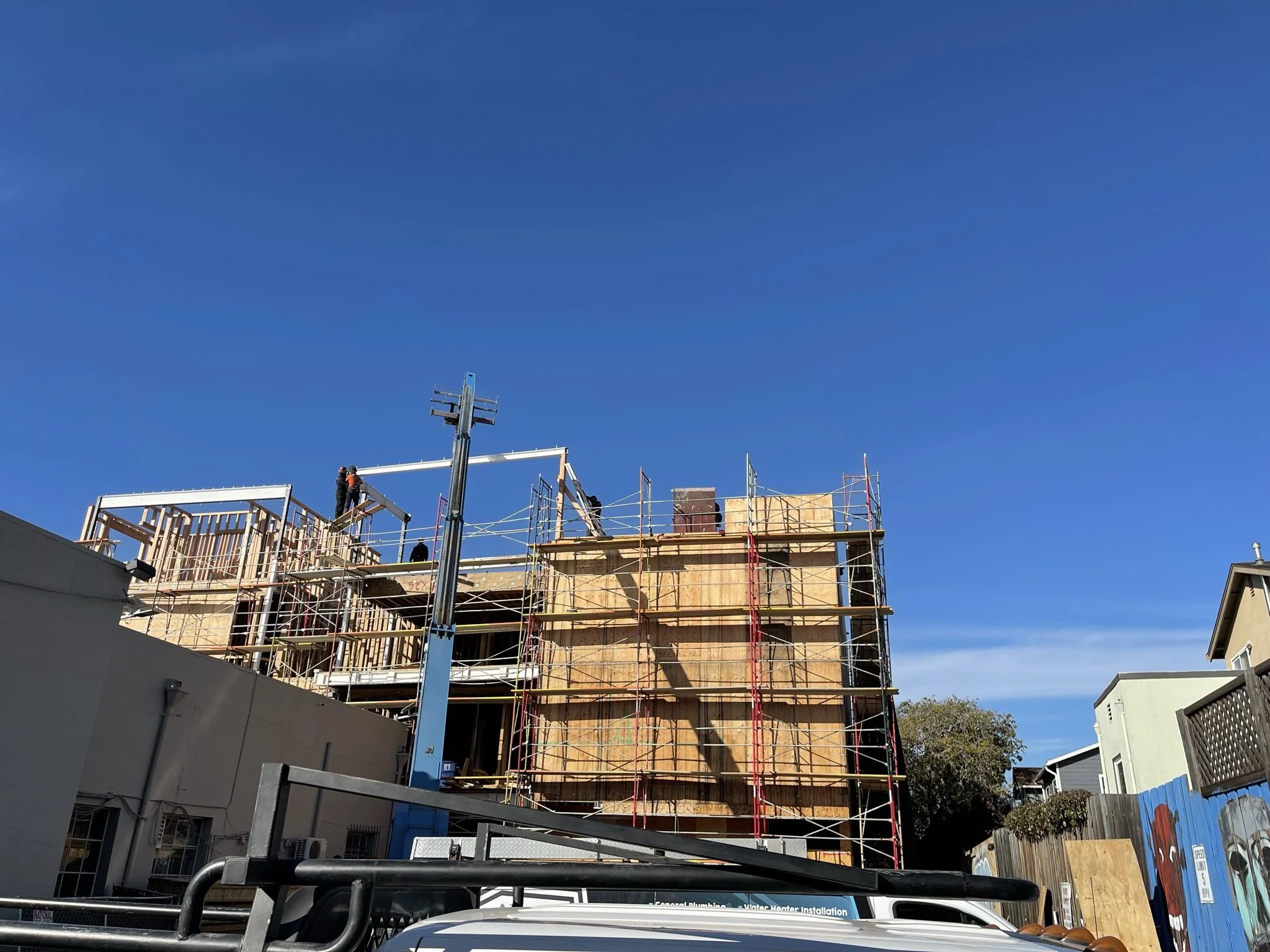 Construction workers building a multi-story wooden structure on a clear day with a blue sky, surrounded by scaffolding.