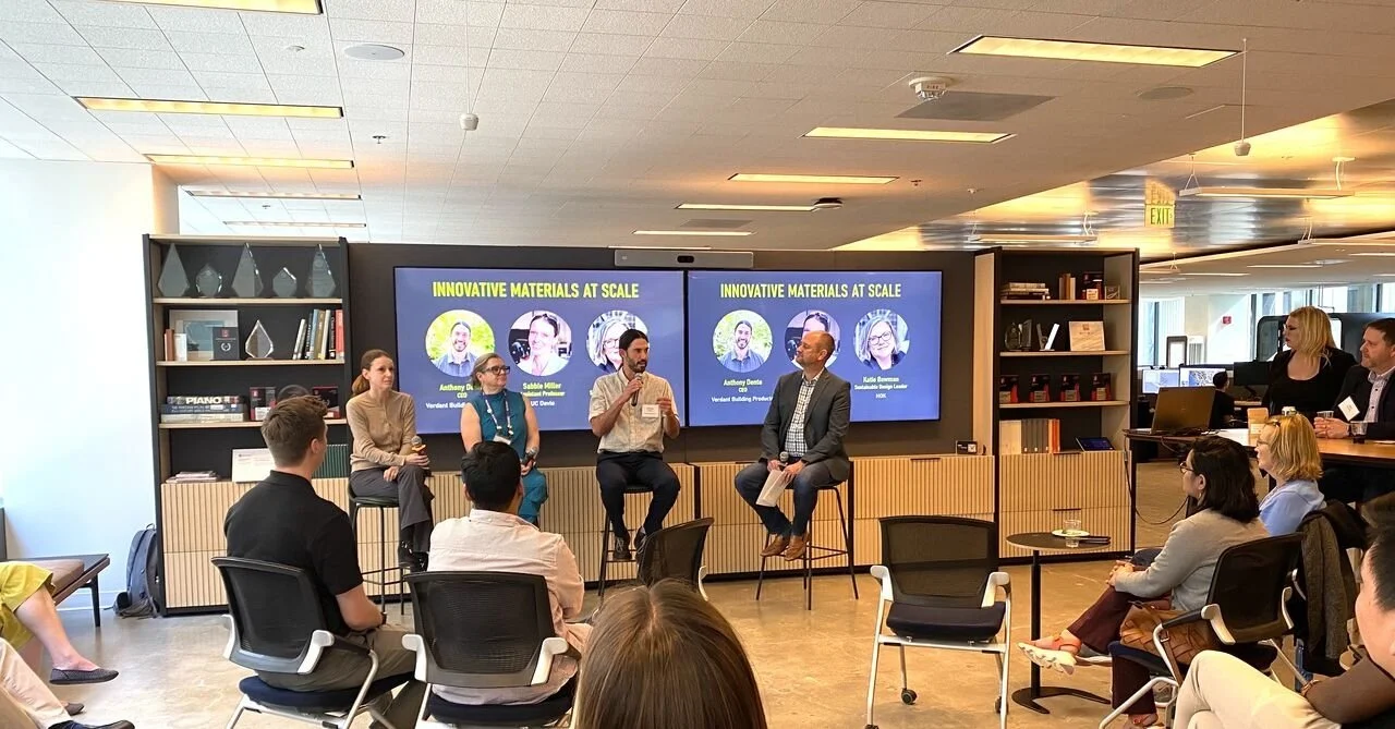 A panel discussion with four speakers seated on stools in front of a screen displaying 'Innovative Materials at Scale' and images of three speakers. Audience members sit and listen in a modern conference room.