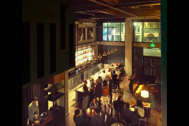 Inside a modern restaurant with people ordering at the counter and sitting at tables, illuminated by warm lighting and large window view of city street.