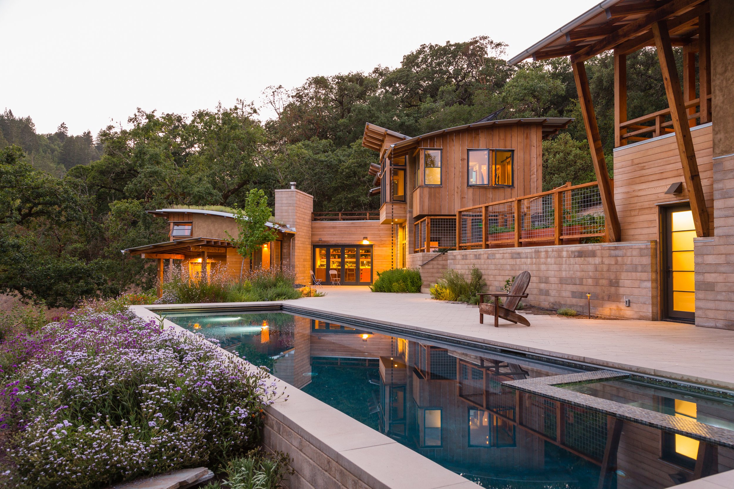 Modern house with large windows and warm lighting, situated in a lush, wooded area at dusk. Features an outdoor swimming pool and a garden with blooming flowers.