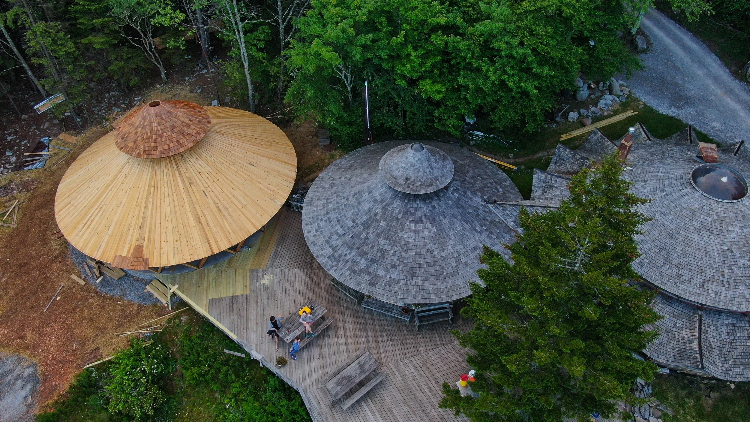 An aerial view with several wooden structures, including two circular turret-like buildings with shingled roofs, a deck area surrounded by tall green trees, with some construction materials and tools nearby.