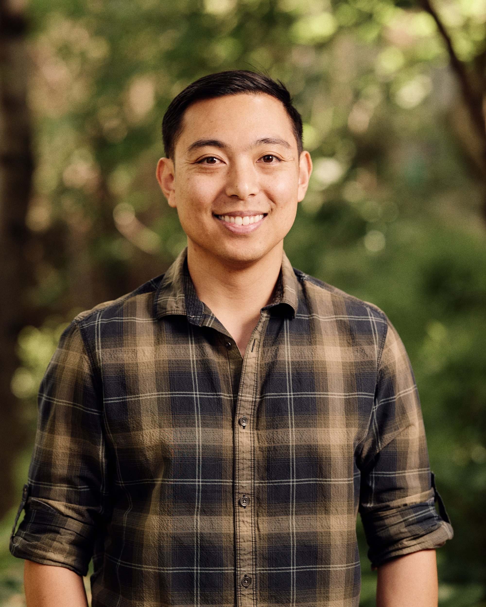 Close-up of a smiling young man with short dark hair wearing a brown plaid shirt, standing outdoors with a blurred green background.