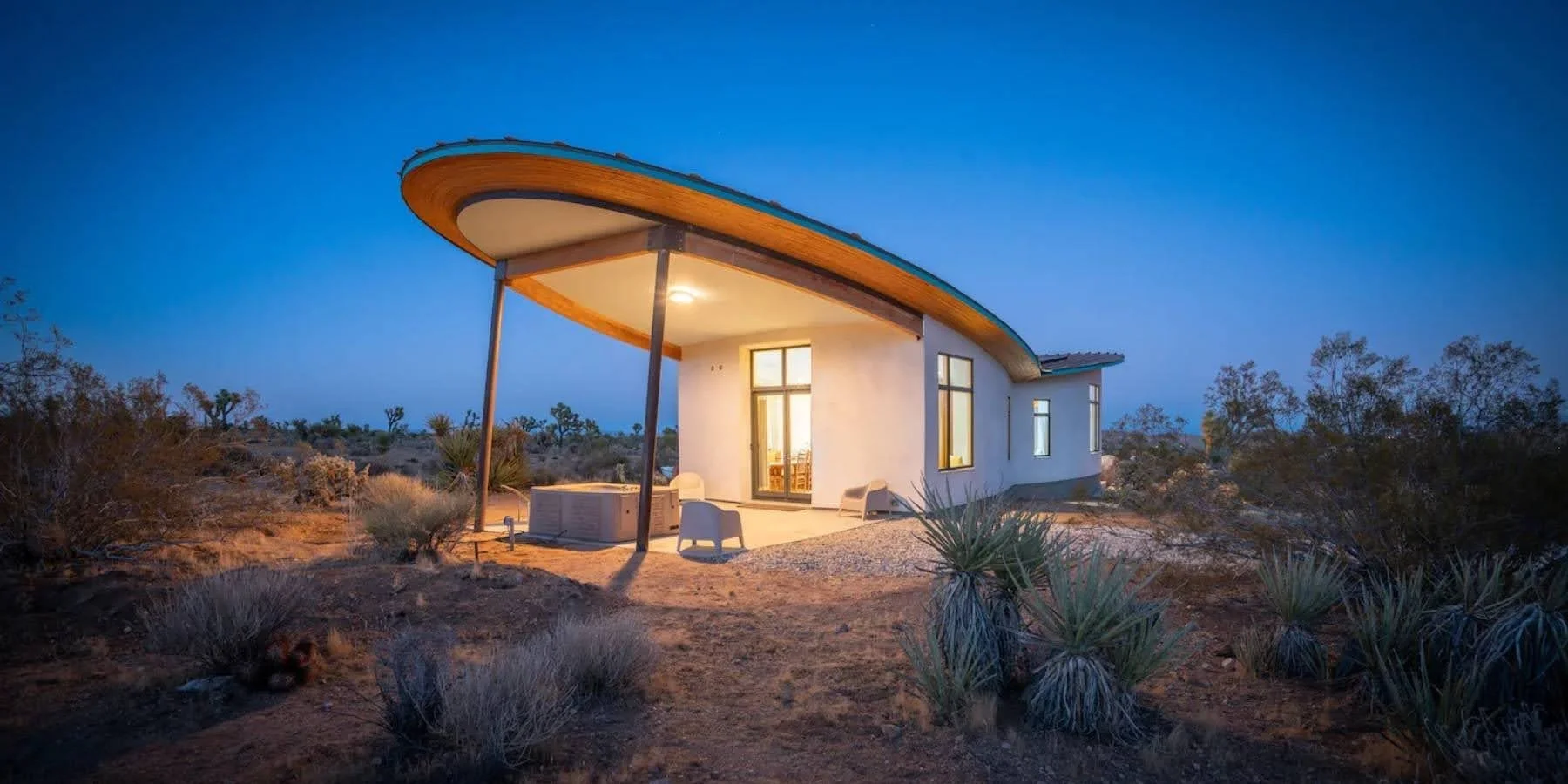 Modern house with curved roof in desert landscape at dusk, surrounded by bushes and plants.