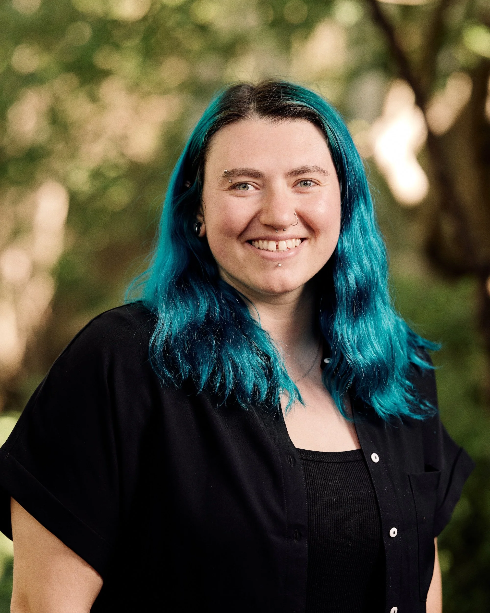 Close-up of young woman with vibrant blue hair, piercing smile, multiple facial piercings, wearing a black top, outdoors with blurred trees in background.