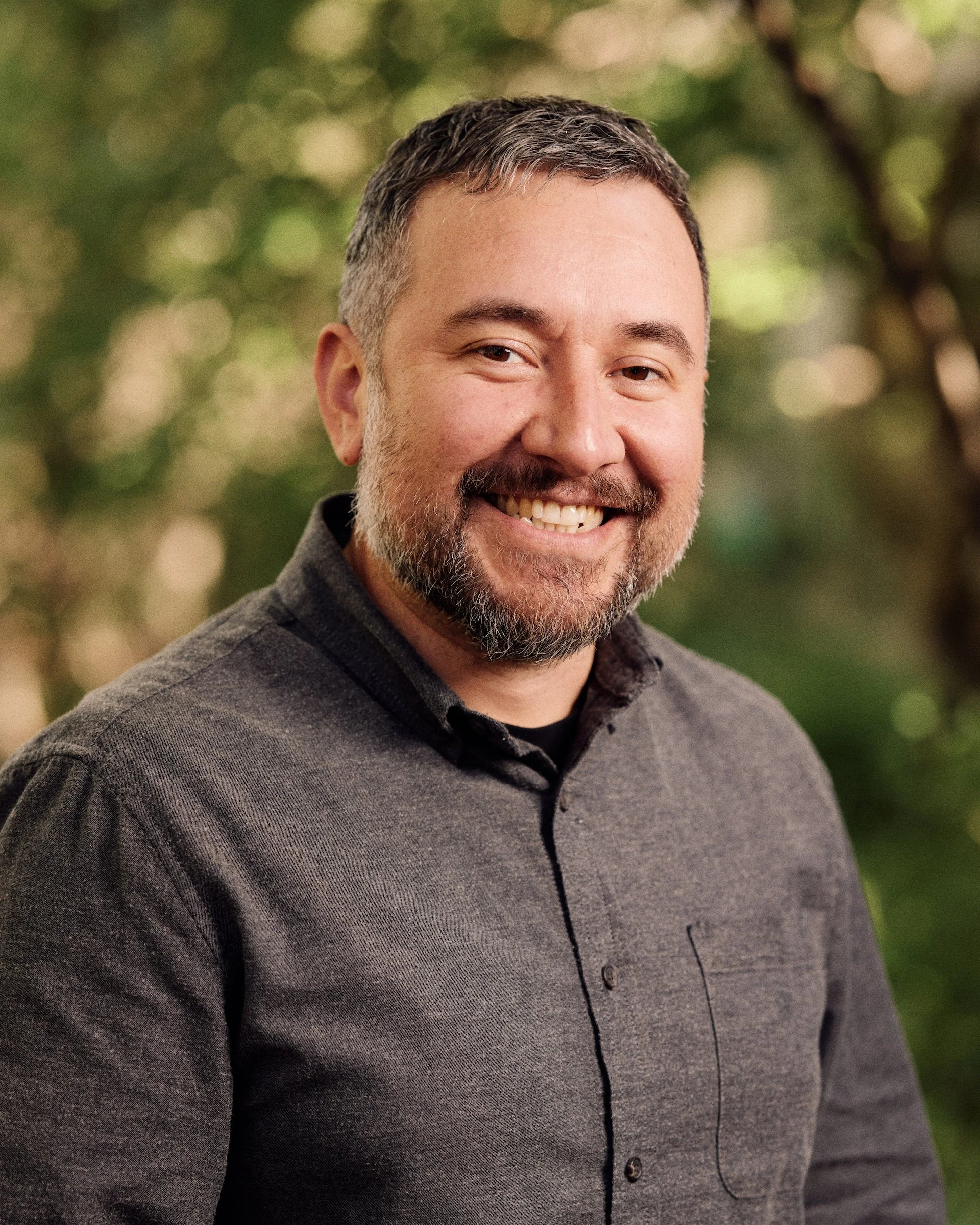 Close-up of a smiling man with short, dark hair, a beard, and mustache, wearing a dark gray shirt, against a blurred green background.