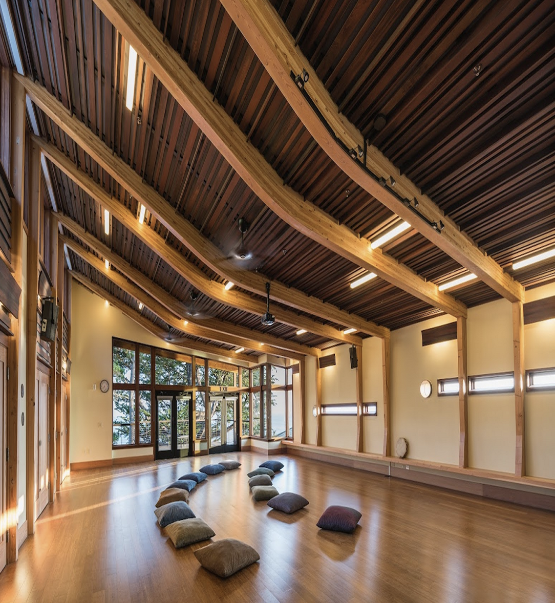 Interior of a wooden meditation or yoga studio with cushions arranged in a circle on a polished wood floor, large windows, and modern lighting.