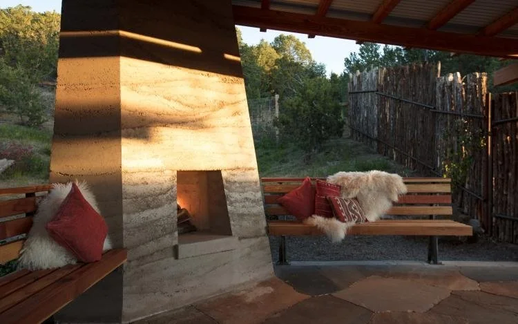 Outdoor seating area with a large stone fireplace, wooden benches with red pillows and sheepskin throws, surrounded by a wooden fence and trees in the background.