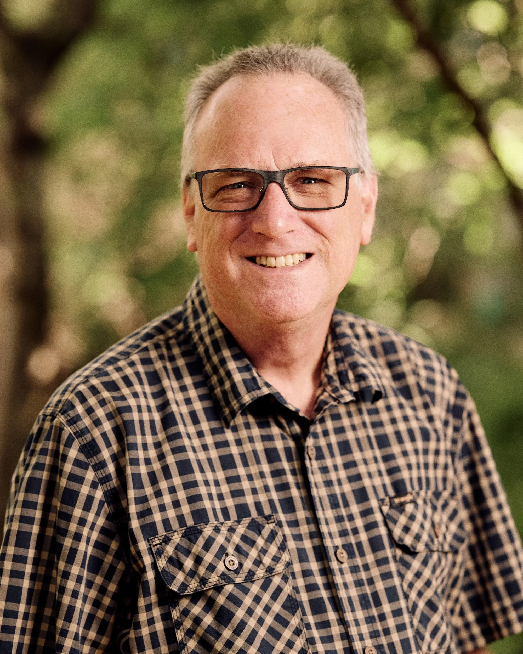 Close-up of a smiling middle-aged man with gray hair, wearing black-rimmed glasses and a checkered shirt, standing outdoors with blurred green foliage in the background.