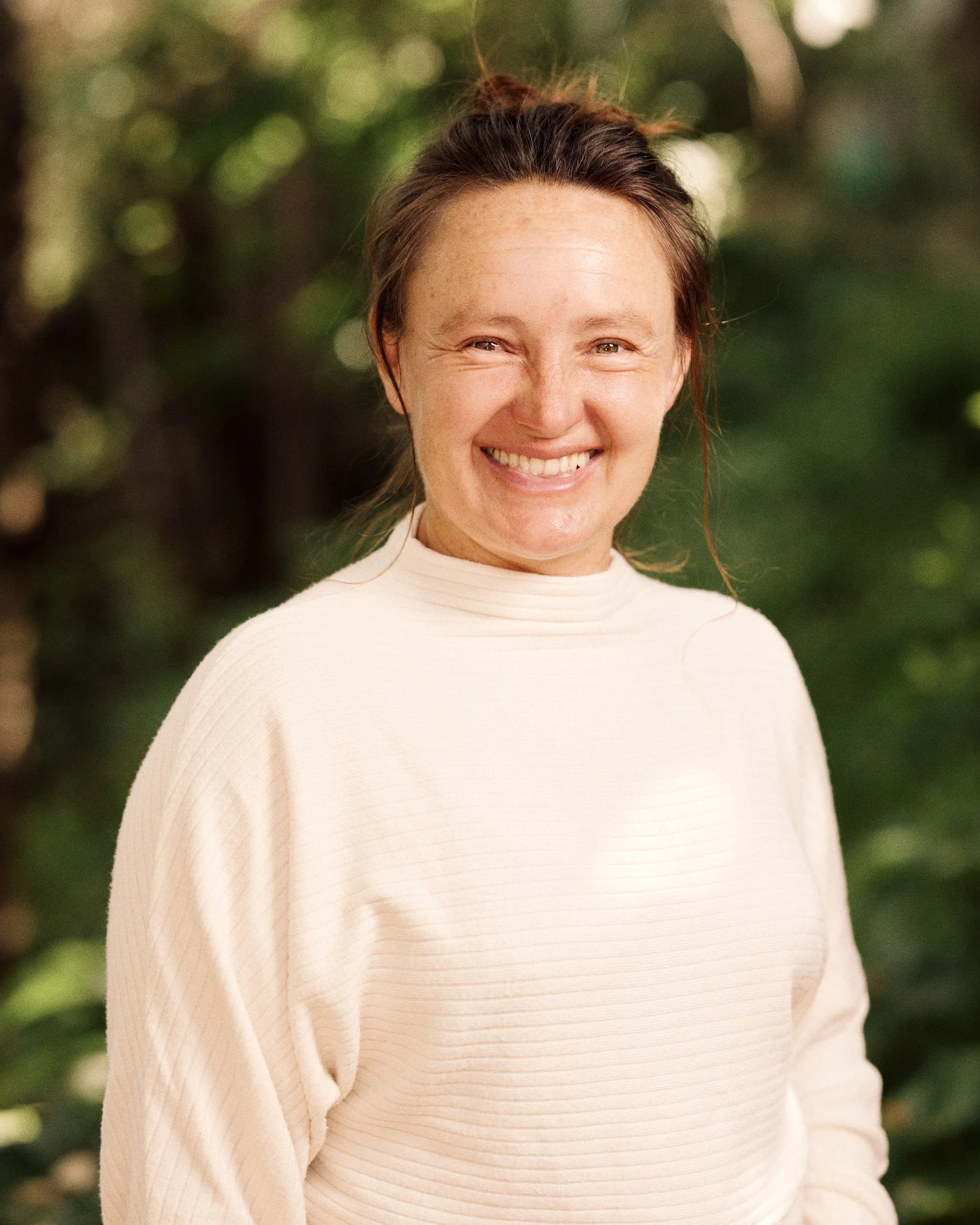 A woman smiling outdoors with blurred green foliage background, wearing a cream-colored sweater.