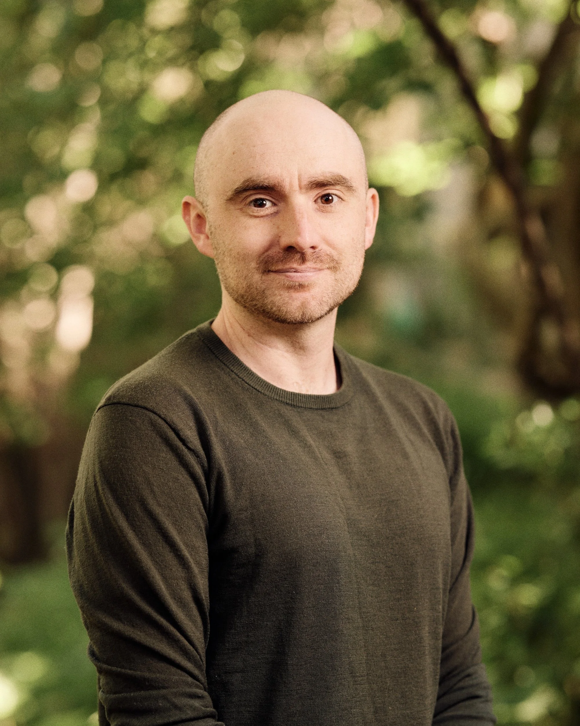 A man with a shaved head and facial hair standing outdoors with trees and greenery in the background.