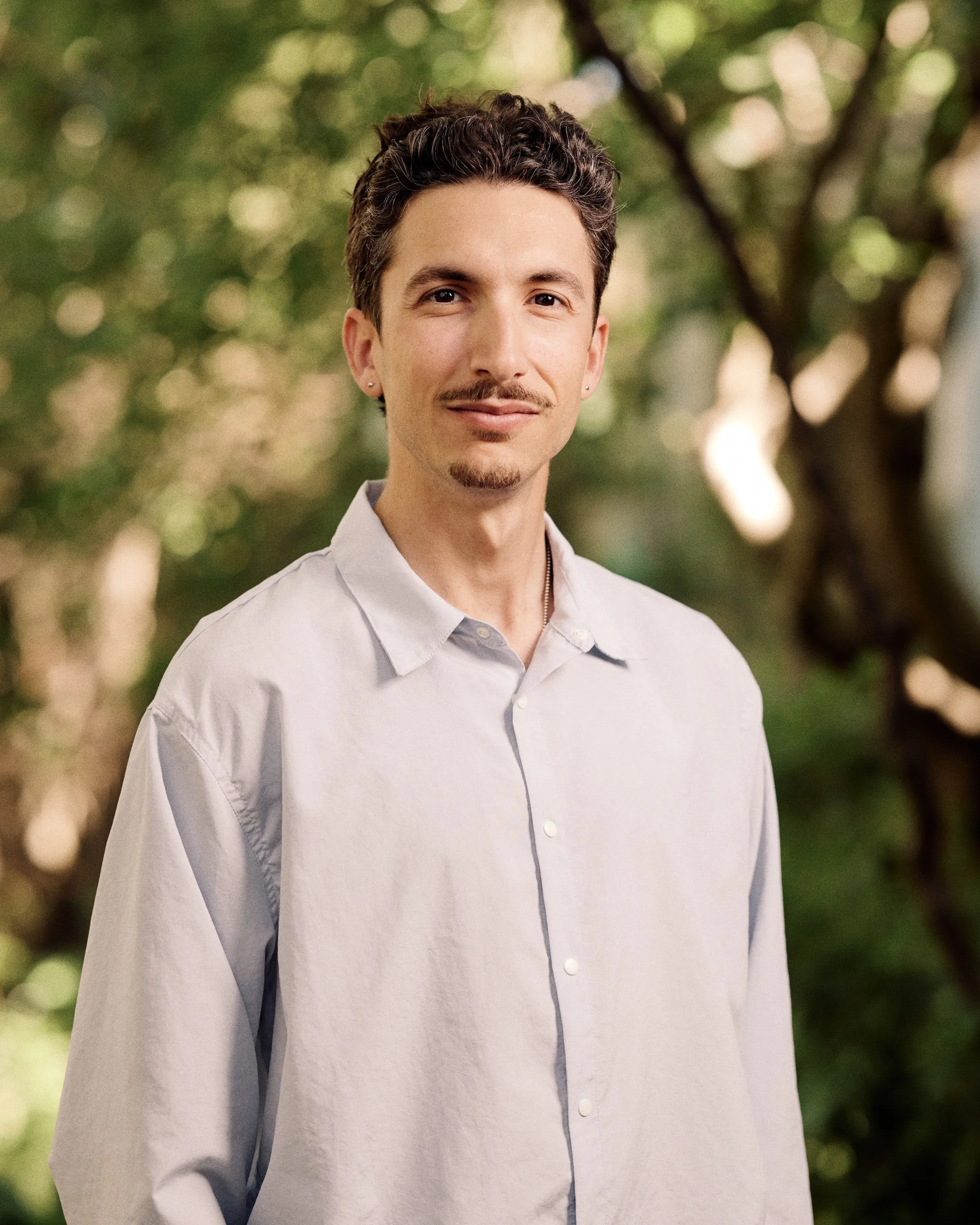 Close-up of a young man with dark curly hair, light skin, wearing a light-colored button-up shirt and earrings, standing outdoors with a blurred green background of trees.