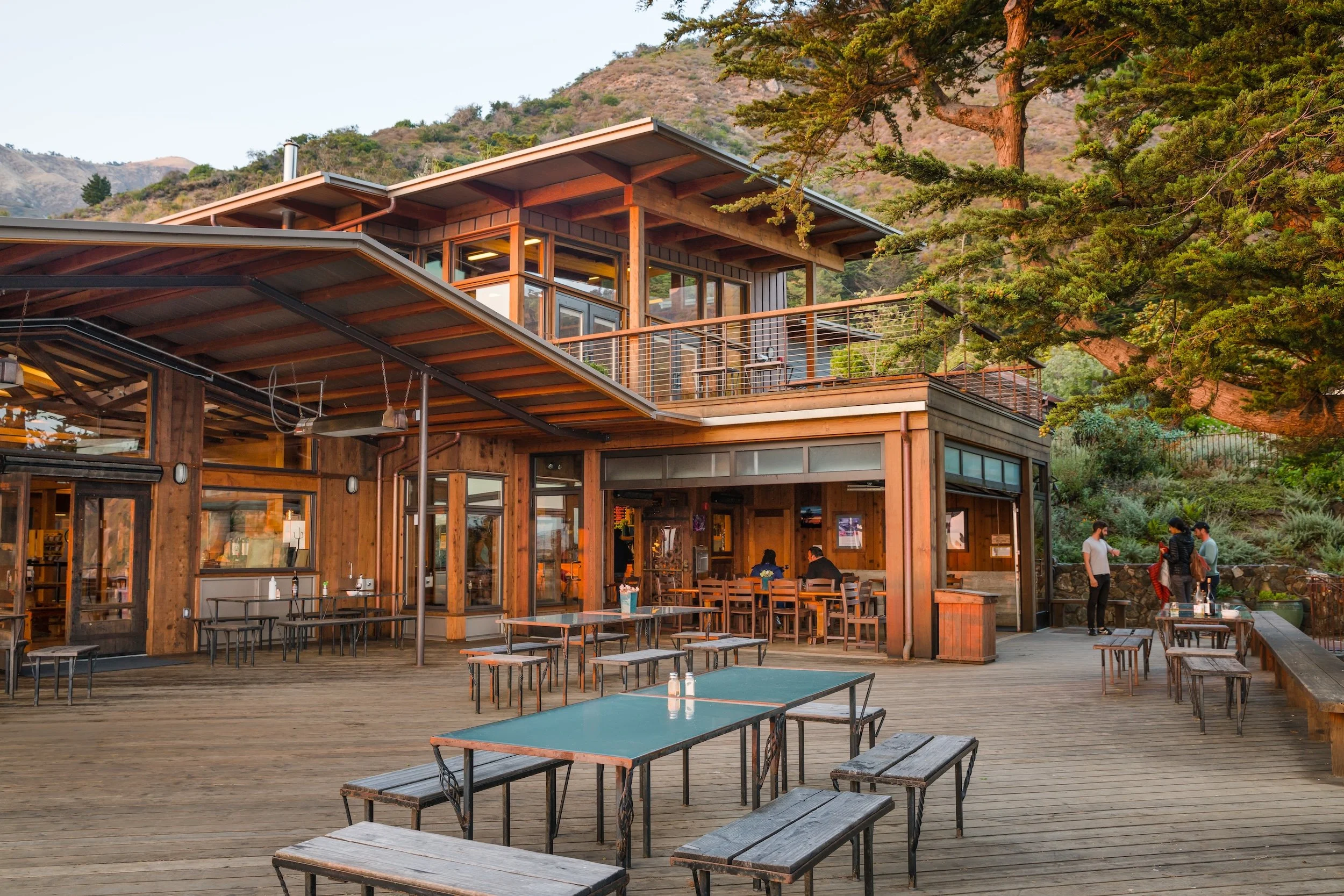 Outdoor patio area at a restaurant or cafe with wooden tables and benches, people dining and standing around, and a modern building with large windows and a deck, surrounded by trees and mountains in the background.
