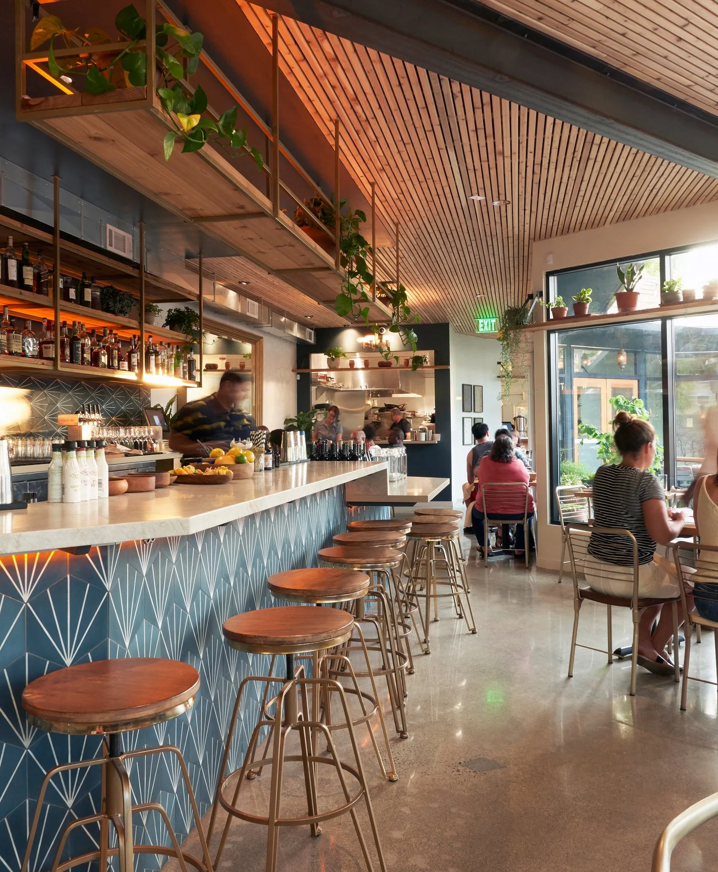 Interior of a modern cafe with a bar counter, barstools, and patrons seated at tables near large windows, decorated with plants and wooden ceiling panels.