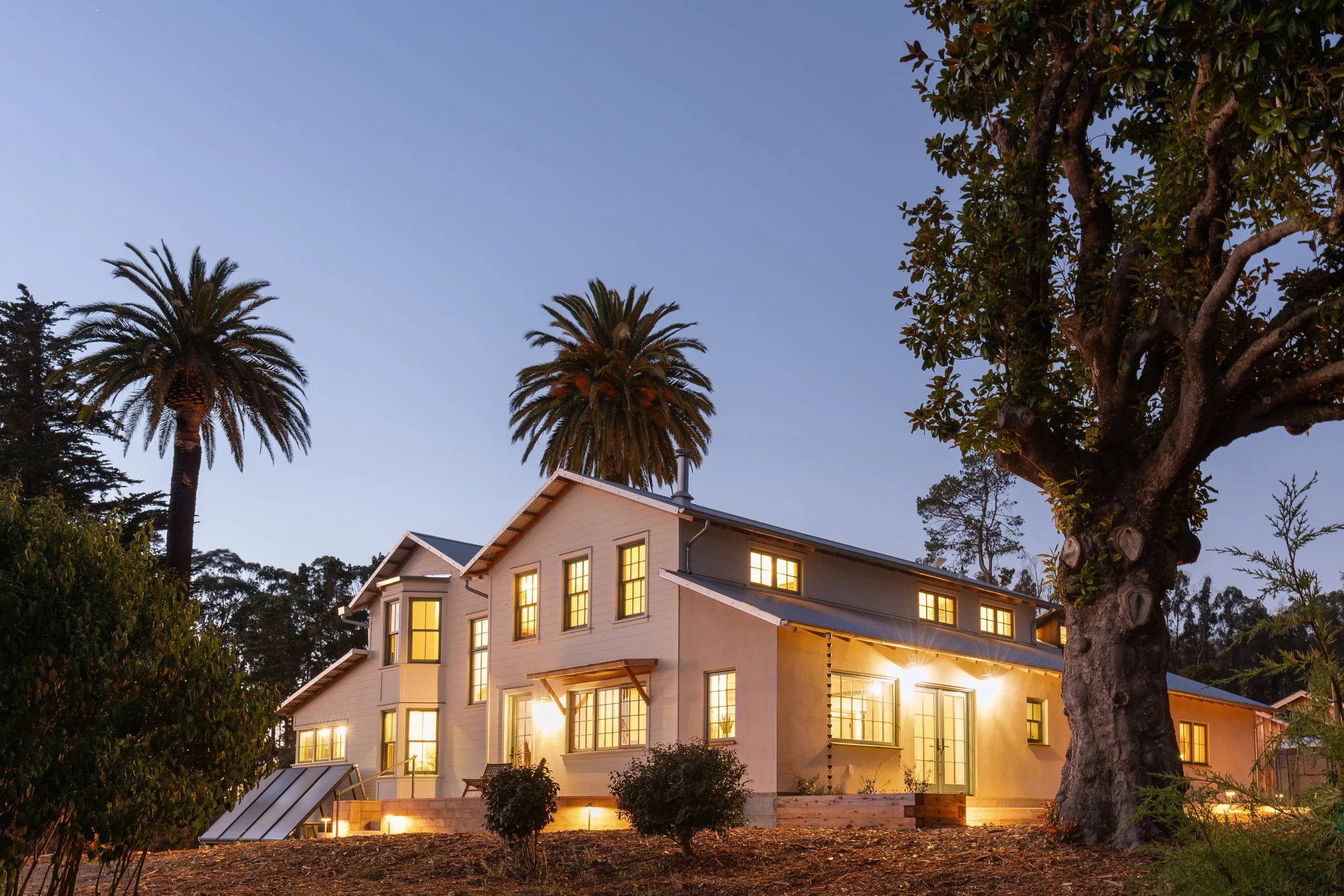 A multi-story house with large illuminated windows, surrounded by trees including palm trees, during dusk or evening.