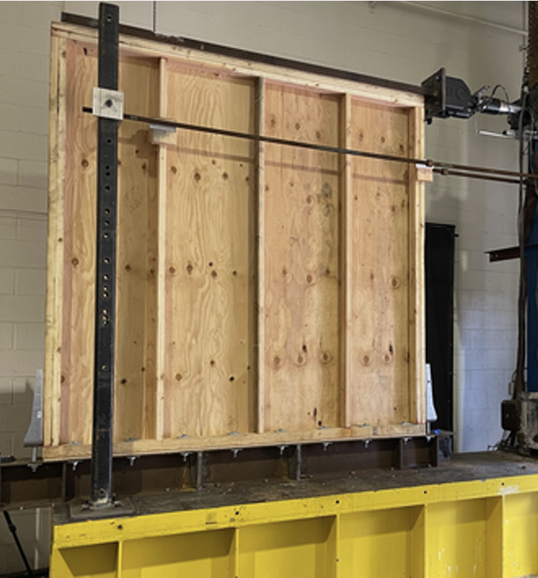 Wooden frame structure with vertical studs, mounted on a yellow work surface in a workshop.