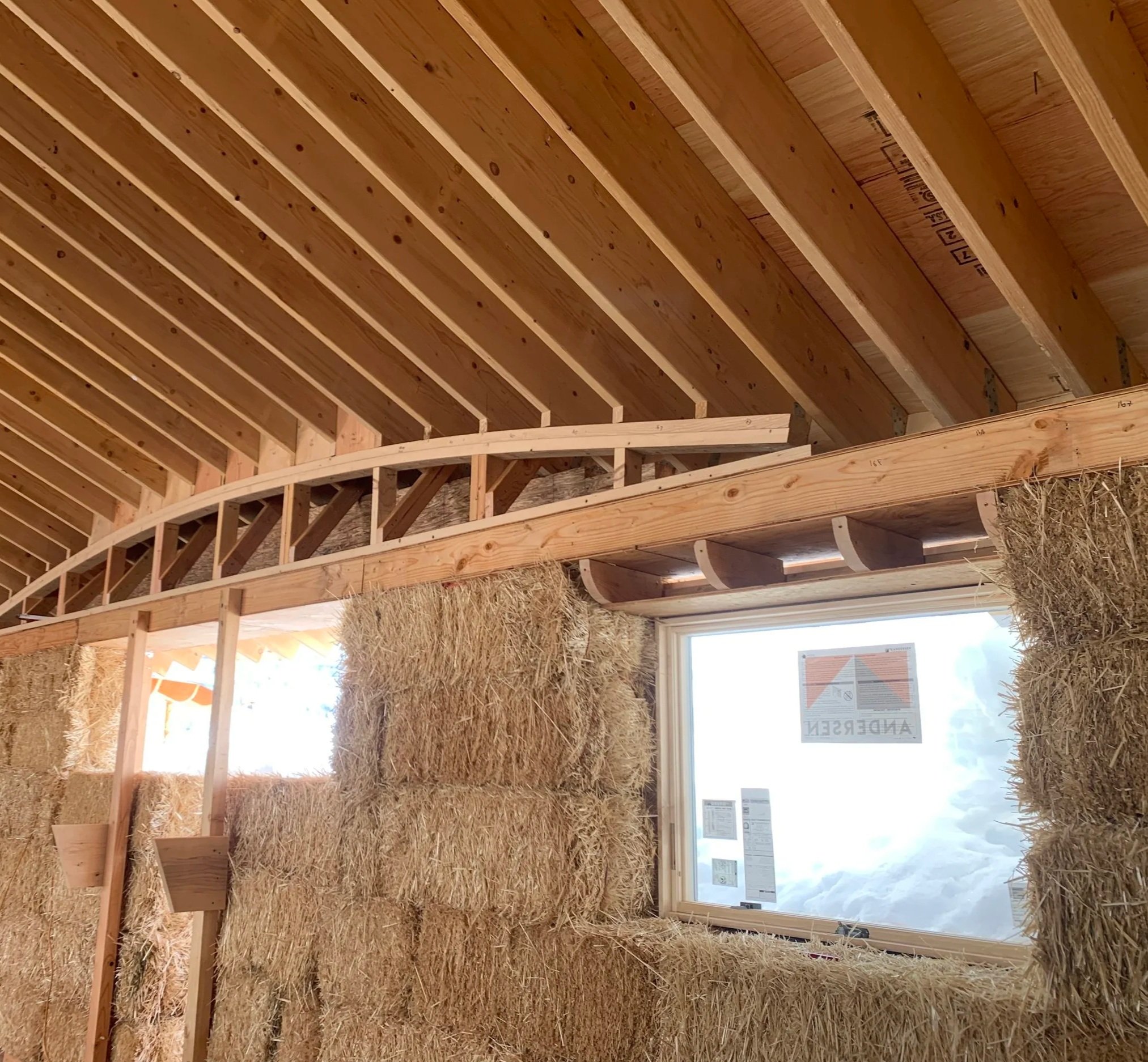 Interior view of a building under construction with wooden framing, straw bales stacked along the wall, and a window with a view of the sky.