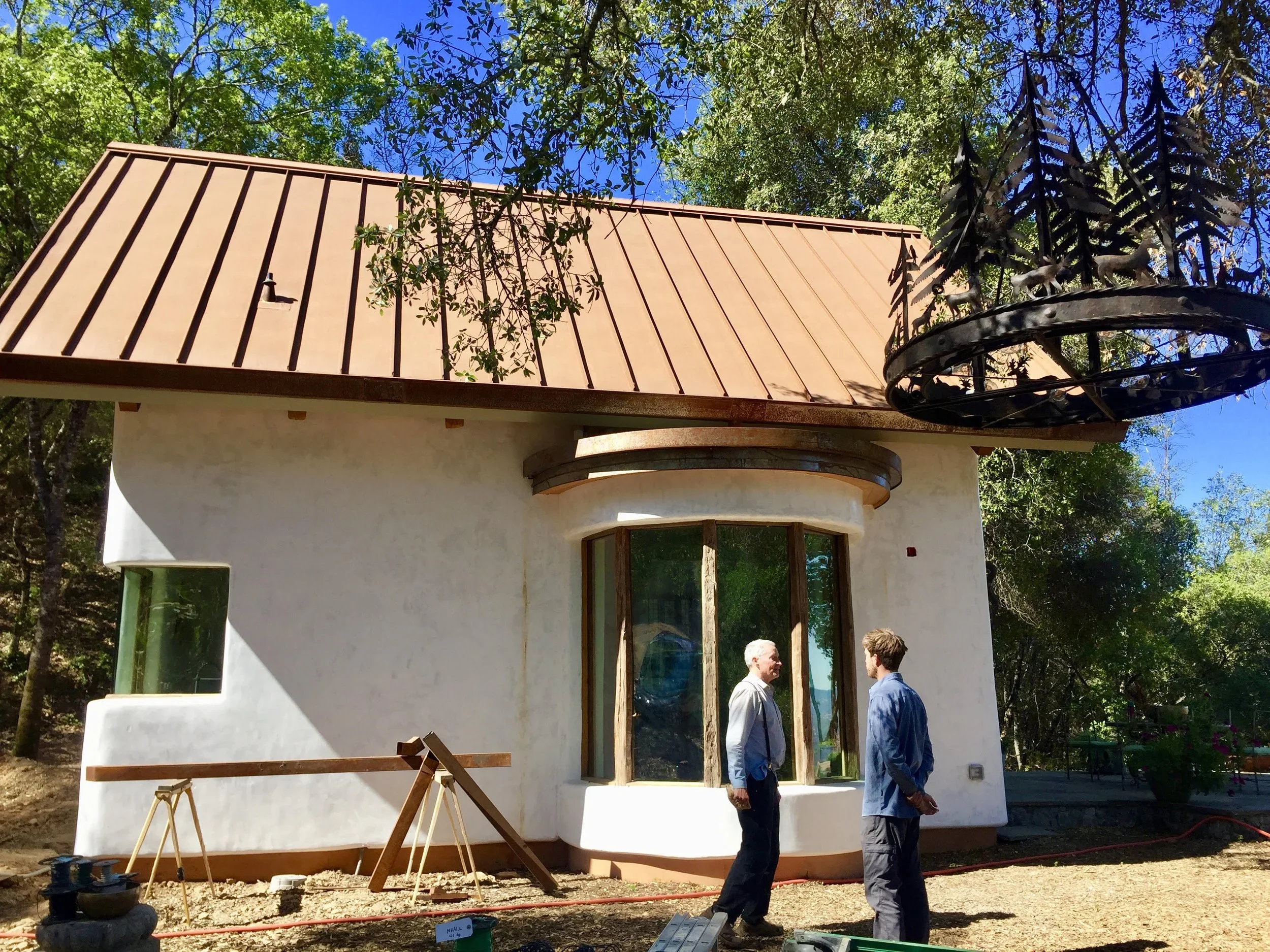 Two men are standing outside near a white building with a copper-colored roof, surrounded by trees. The building appears to be under construction or renovation, with building materials and tools visible. A unique chandelier with horse sculptures hangs on the right side of the building.