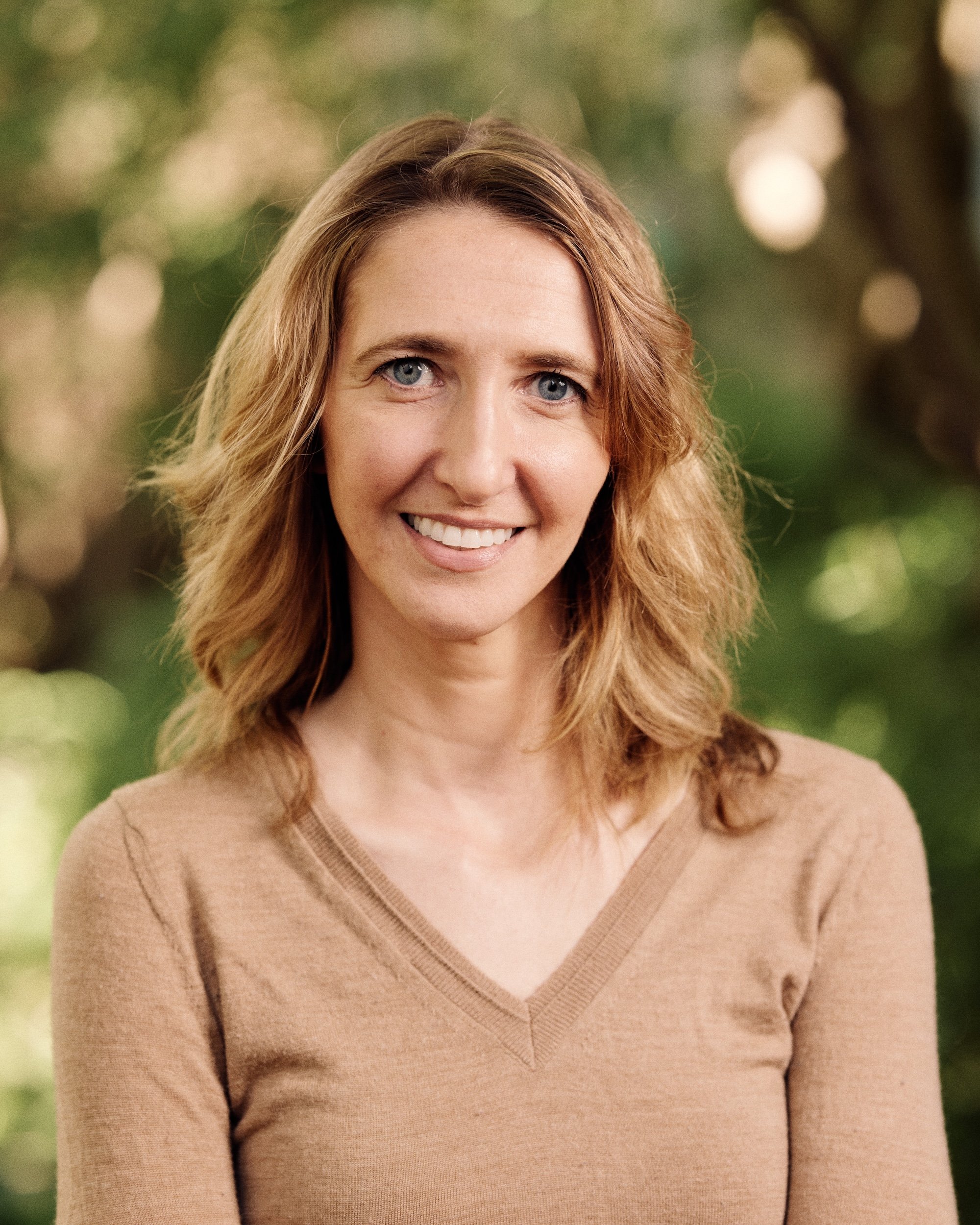 Close-up of a woman with long wavy hair smiling, wearing a beige V-neck sweater, outdoors with blurred green background.