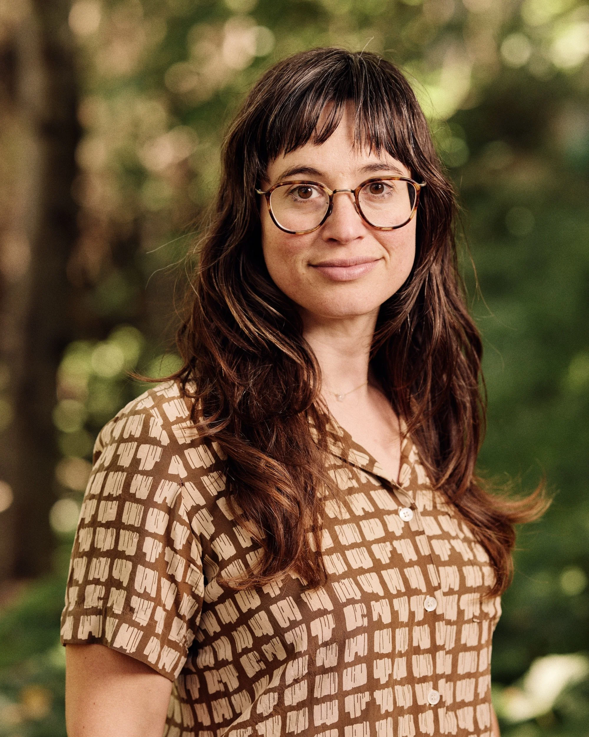 A woman with long, wavy brown hair and glasses, wearing a patterned brown and cream blouse, standing outdoors with blurred green trees in the background.
