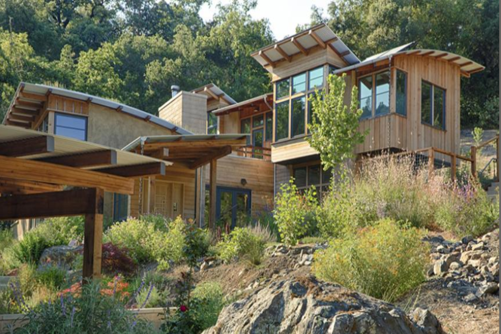 Modern house built on a hillside with large windows and wooden exterior, surrounded by rocks and lush greenery.