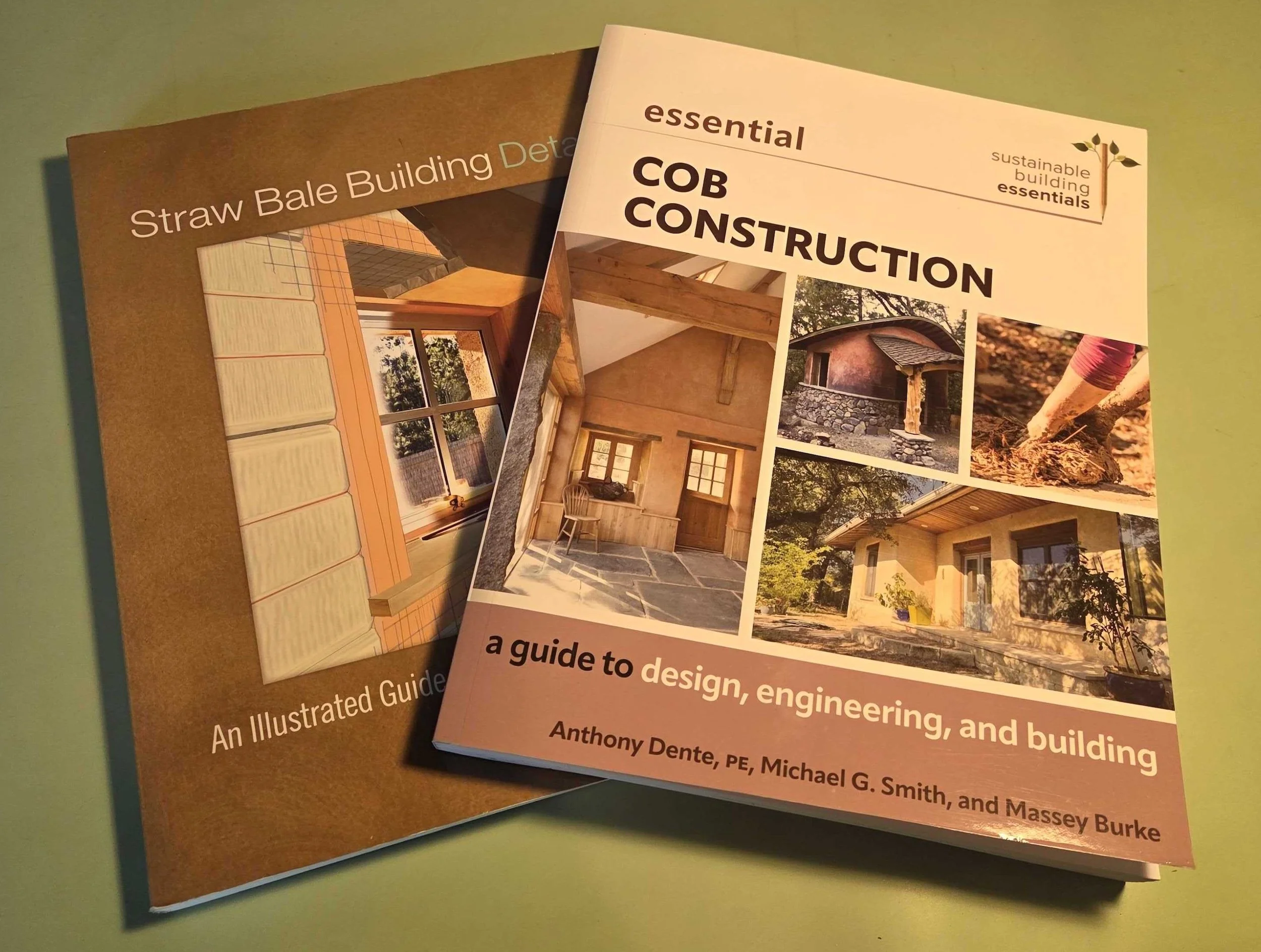 Two books on sustainable building design, one titled "Straw Bale Building Details" and the other "essential COB CONSTRUCTION" with images of wooden structures and construction scenes on the covers.