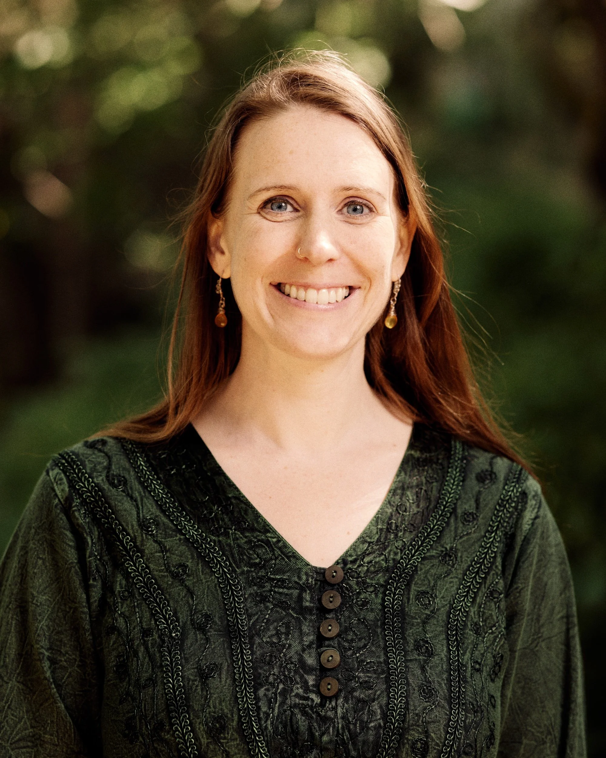 Close-up of a woman with long red hair, blue eyes, wearing a dark green patterned top with buttons, and earrings, outdoor background.
