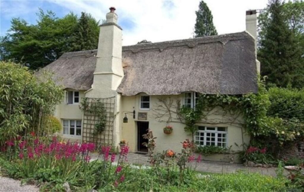 A charming cottage with a thatched roof, white walls, surrounded by lush greenery and colorful flowers.