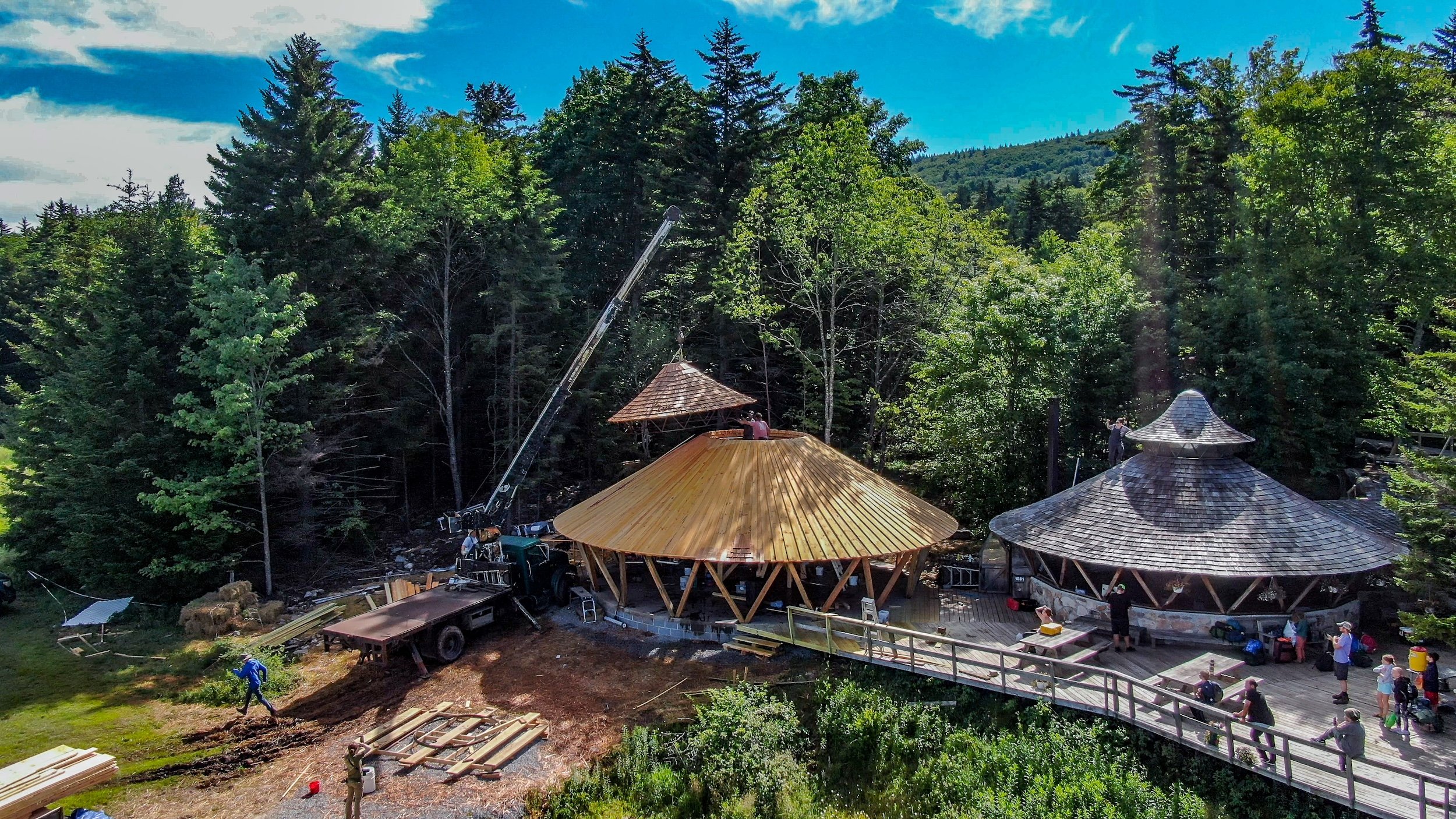 Construction site featuring large wooden domed structures in a forested area with tall trees, partially cloudy sky, workers, and construction equipment.