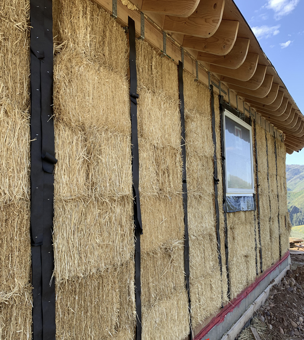 The exterior of a house under construction with straw bales as insulation covered by black straps, and a window with plastic covering, with a blue sky and some clouds in the background.