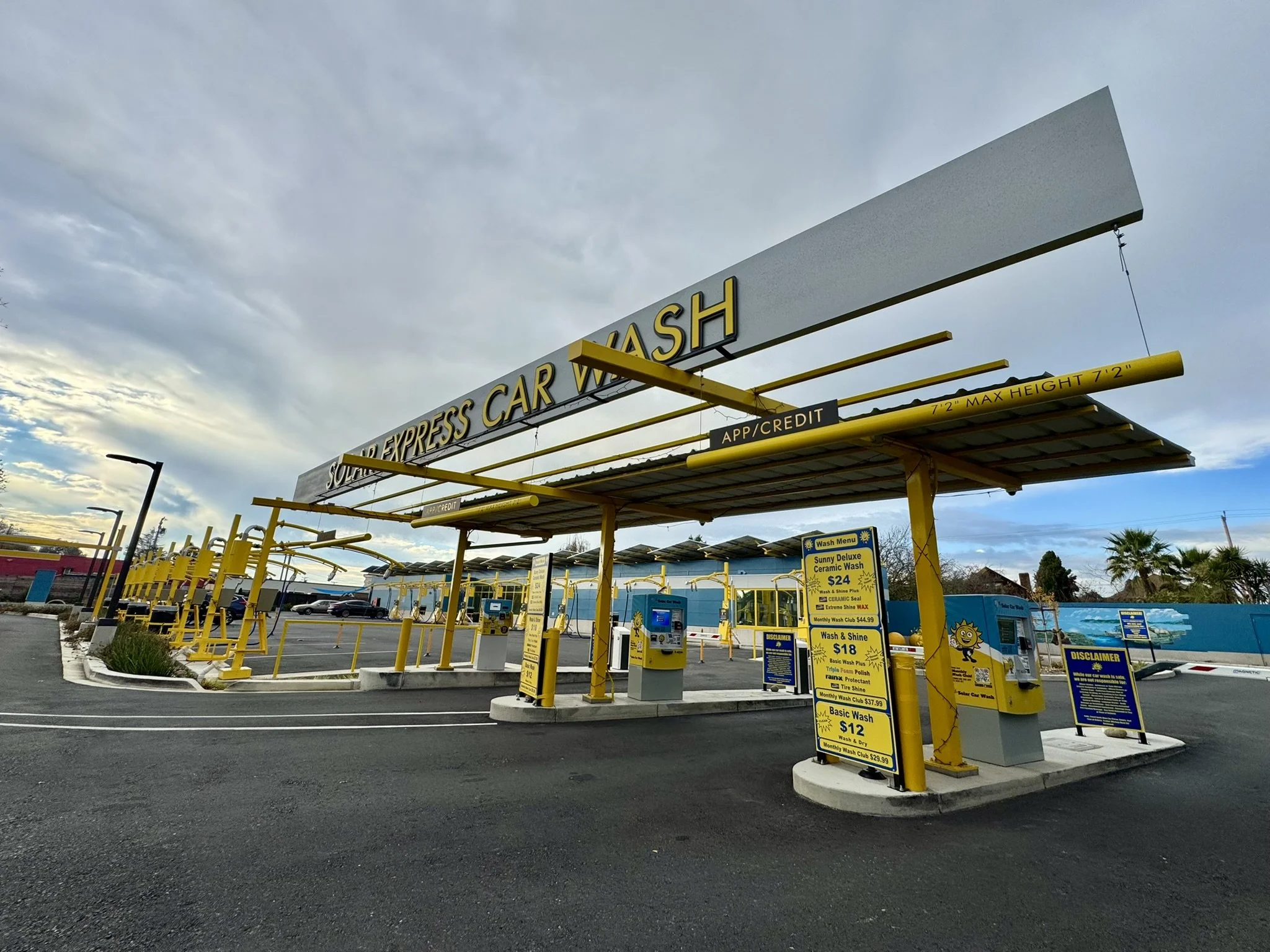 A self-service car wash with yellow structures, signage showing prices for various wash packages, and a partly cloudy sky overhead.