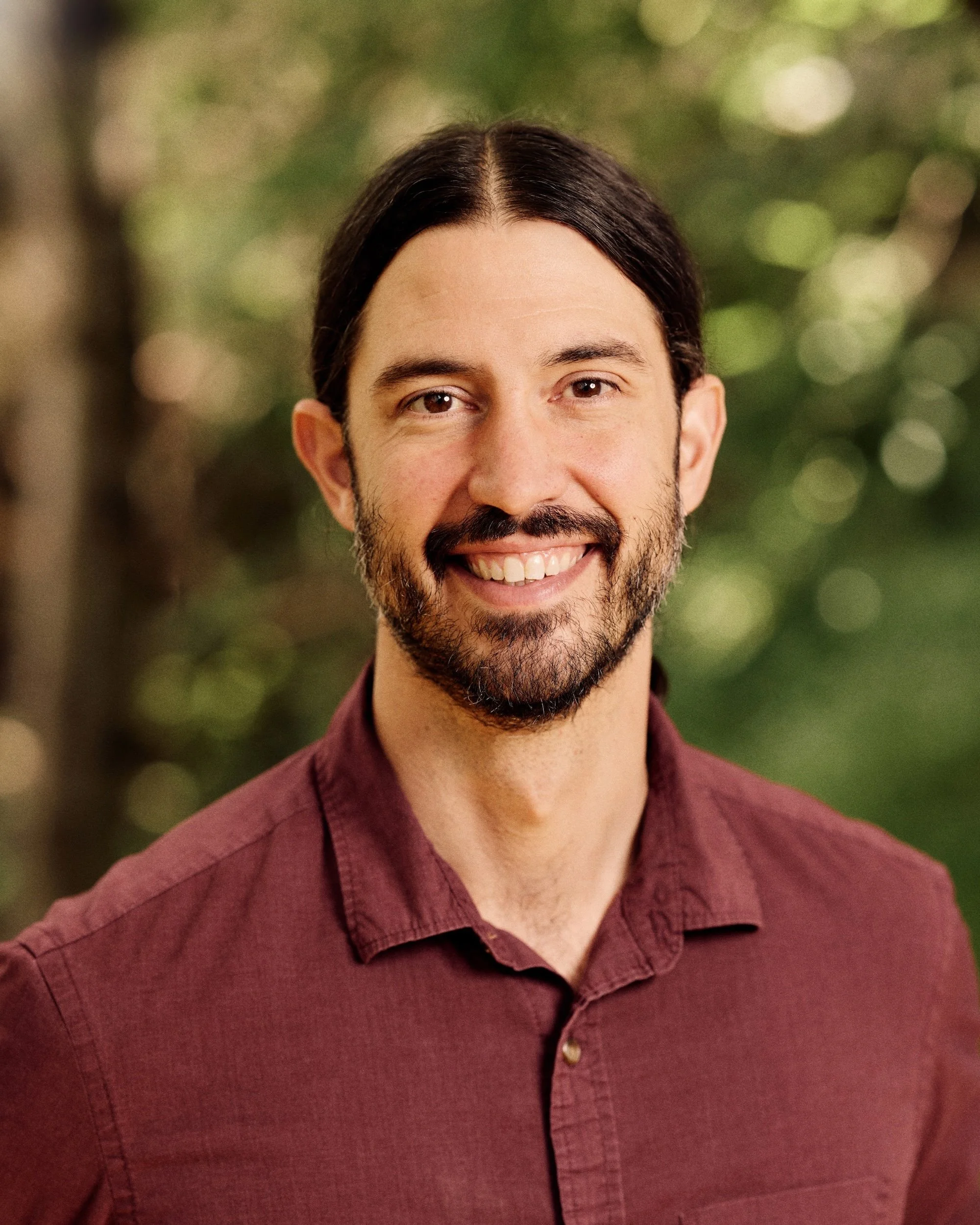 A smiling man with dark hair, a beard, and wearing a maroon button-up shirt. The background is blurred with green and yellow tones.