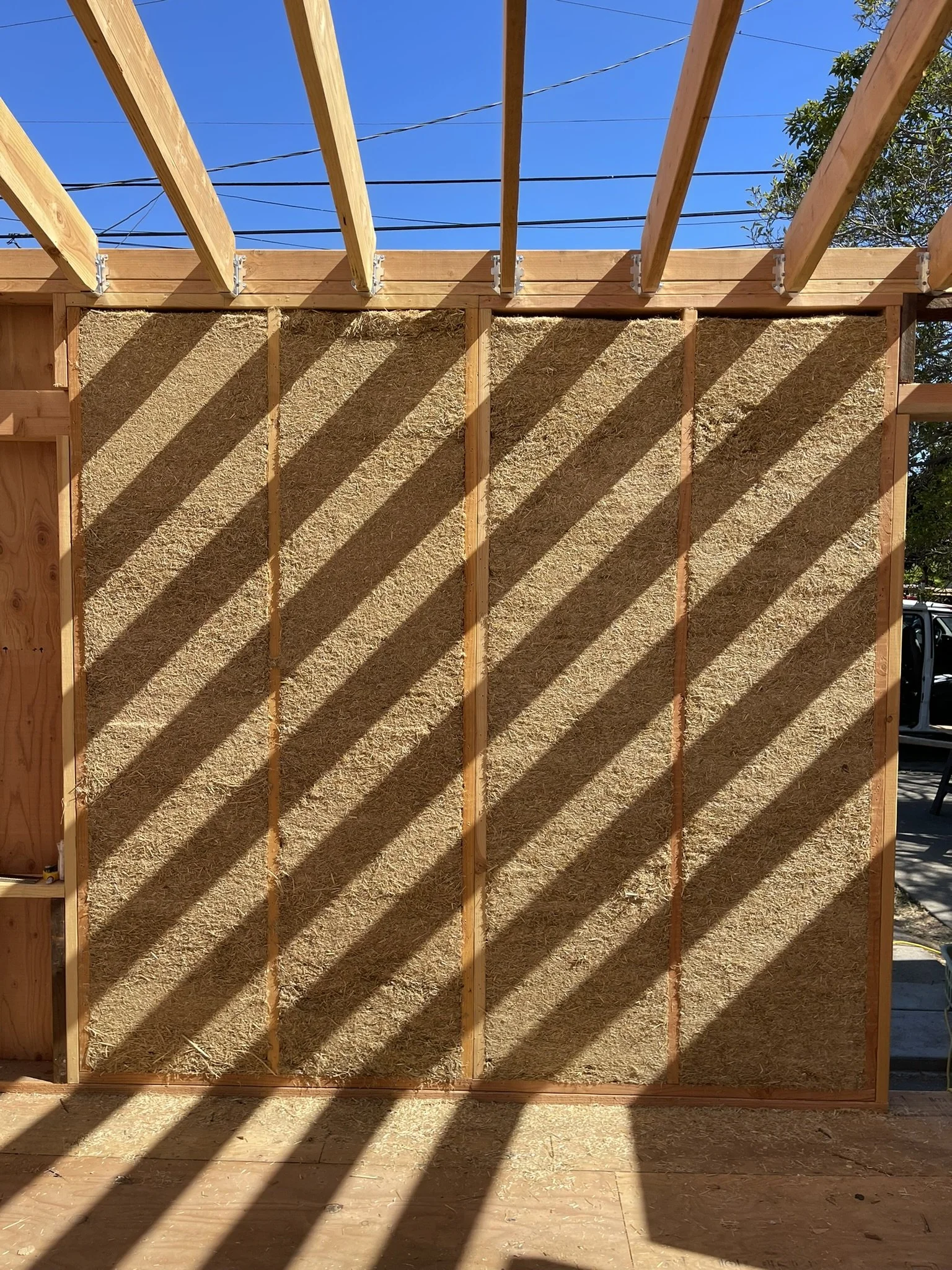 View of a wooden structure under construction with framing and insulation panels, outdoor blue sky, and shadows cast by the wooden beams.
