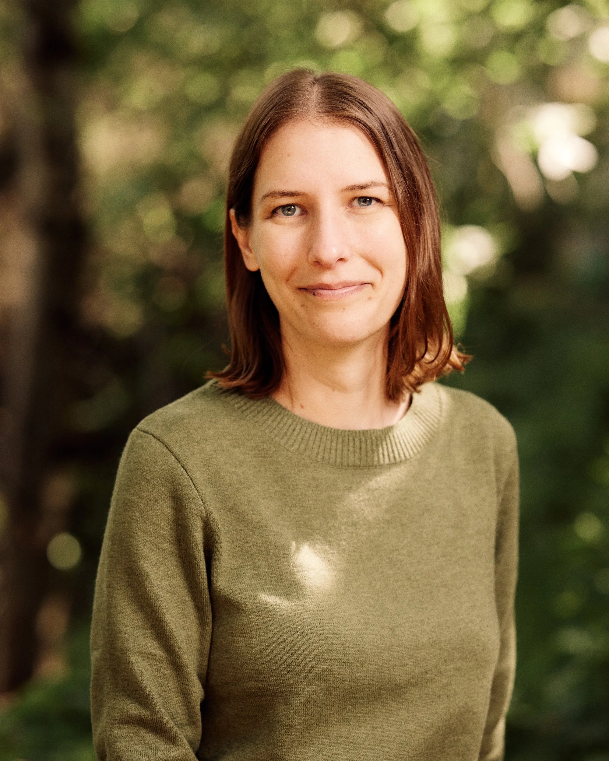A woman with shoulder-length brown hair and light skin smiling outdoors. She is wearing a green sweater, and the background is blurred with trees and sunlight.