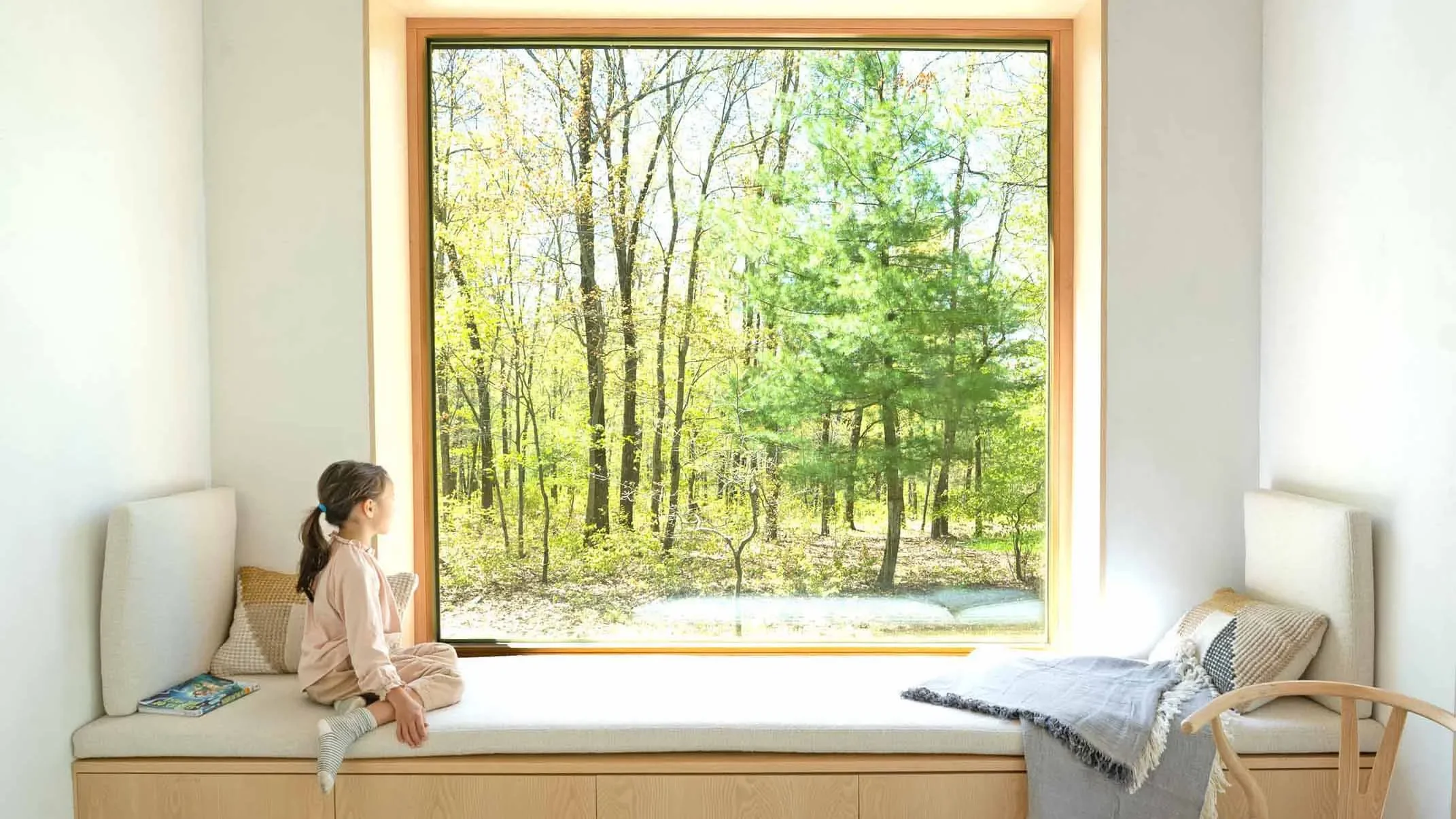 A young girl is sitting on a window seat inside a bright room with white walls. She is looking out at a large window framed in wood, showing a view of a wooded forest with green trees and sunlight.
