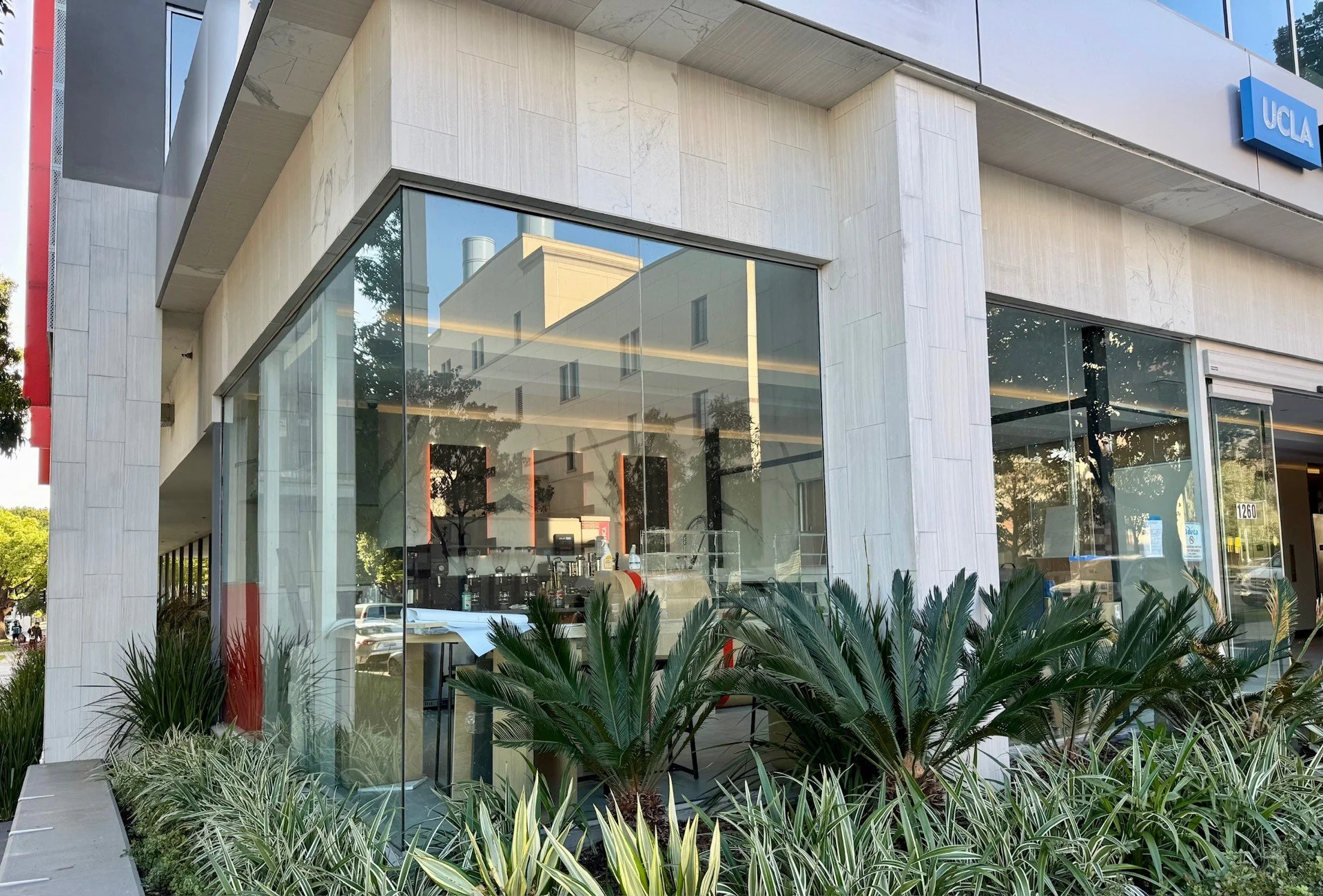 Exterior view of a UCLA building with large glass windows, plants in front, and a reflection of nearby buildings and trees.