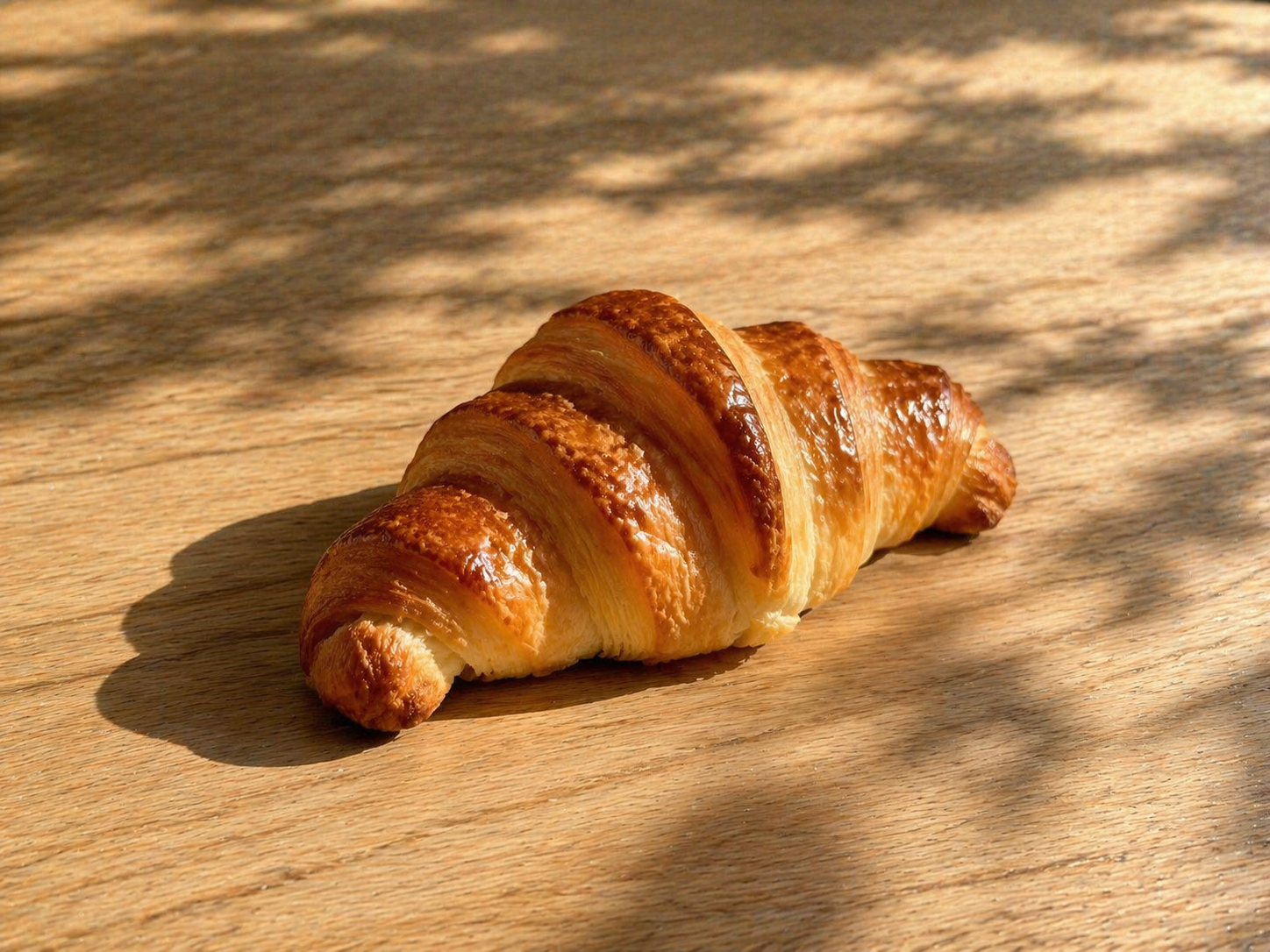 Golden brown croissant on a wooden surface with sunlight and shadows.