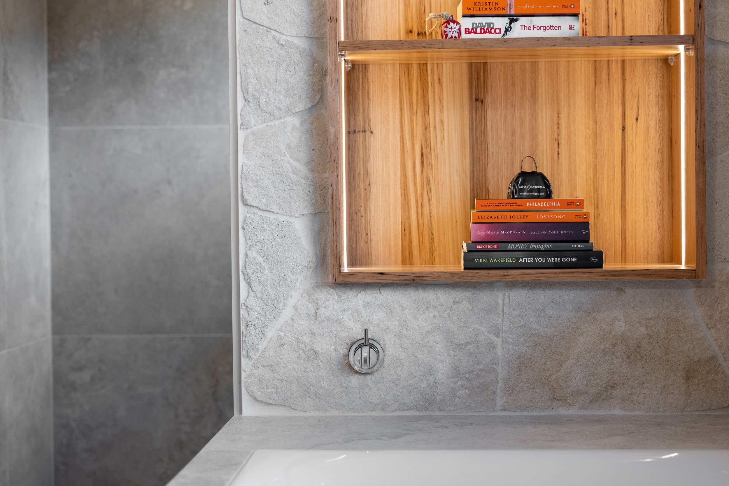 Bathtub with gray stone wall tiles, a wooden shelf with books, and a black alarm clock.