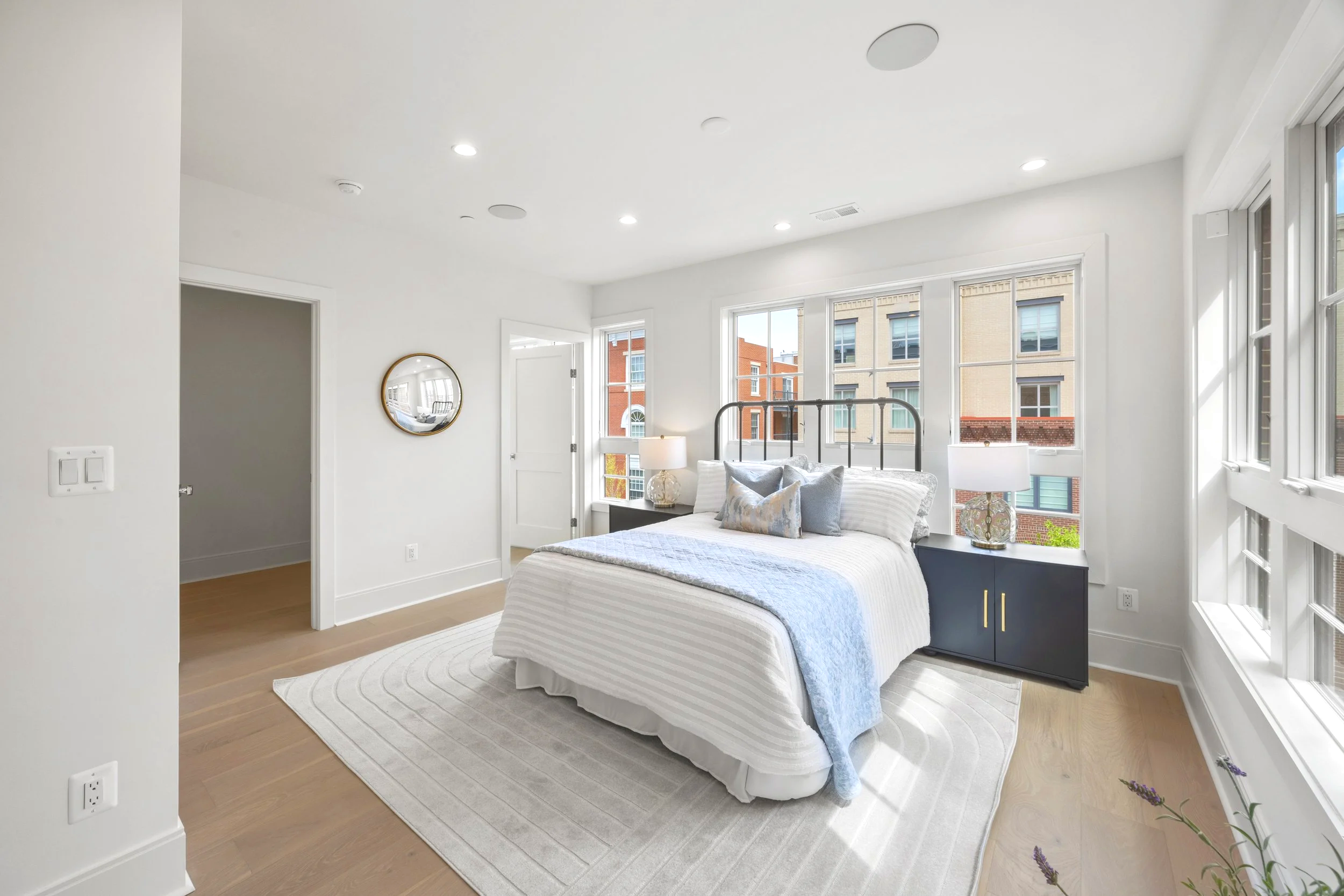Bright bedroom with large windows, a bed with white linens and decorative pillows, two side tables with lamps, and a wall mirror.