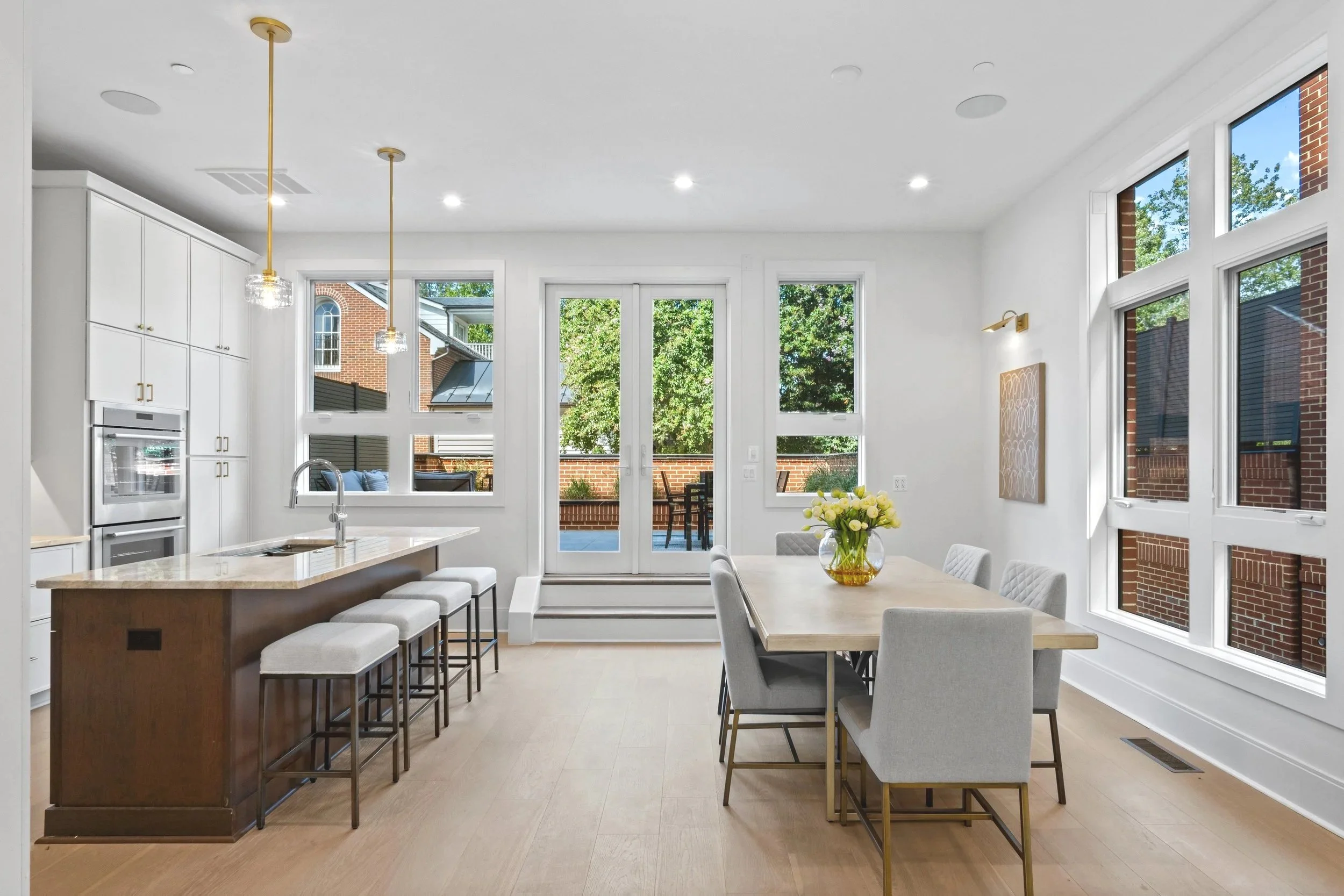 Bright, modern kitchen and dining area with large windows, white walls, and light wood flooring. There is a kitchen island with barstools, a dining table with chairs, and a vase with yellow flowers. Outside, a patio and trees are visible.